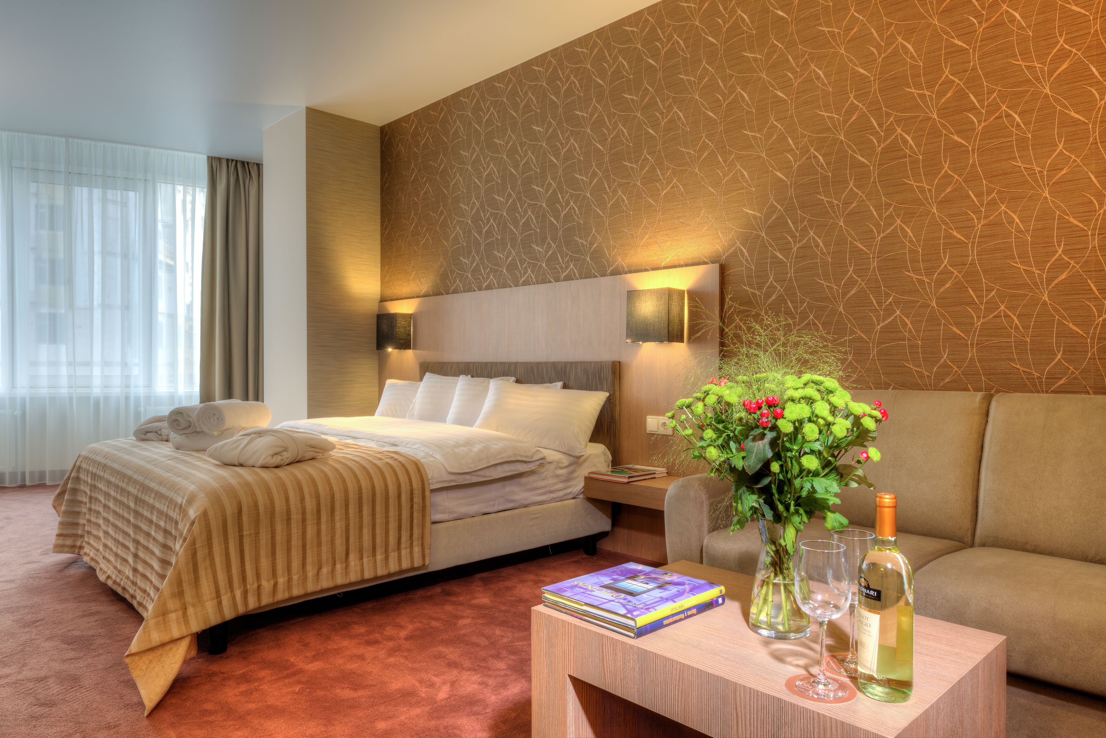Quarto de hotel com cama, sofá, mesa de cabeceira com abajur, mesa com um livro e uma garrafa, e janela com cortinas.