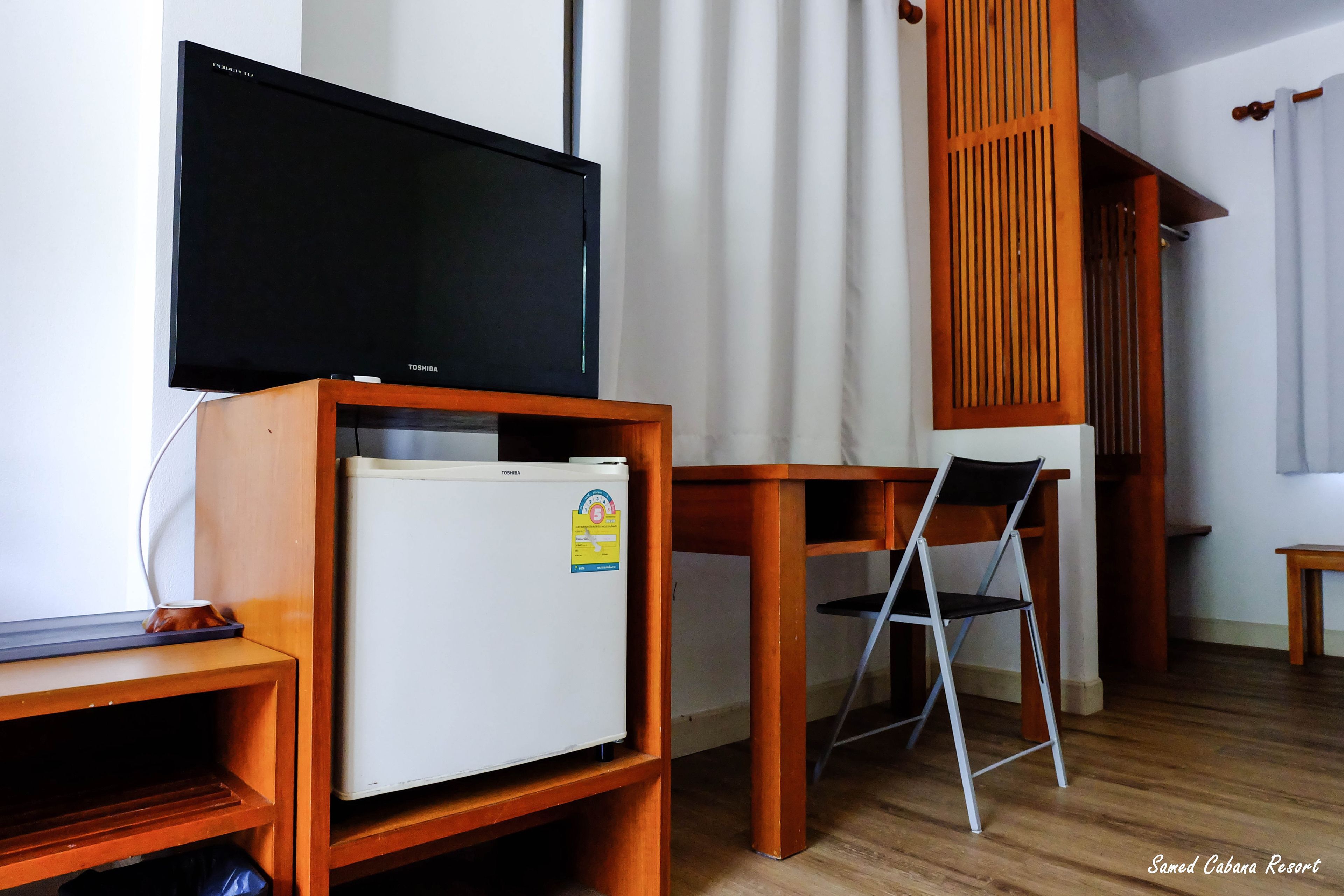 A room with a flat-screen TV on a wooden stand, a small white refrigerator, a foldable chair, and a wooden table.
