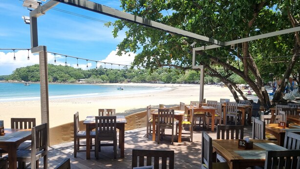 An outdoor seating area with wooden tables and chairs, overlooking a beach and ocean. - Samed Cabana Resort (Rayong)
