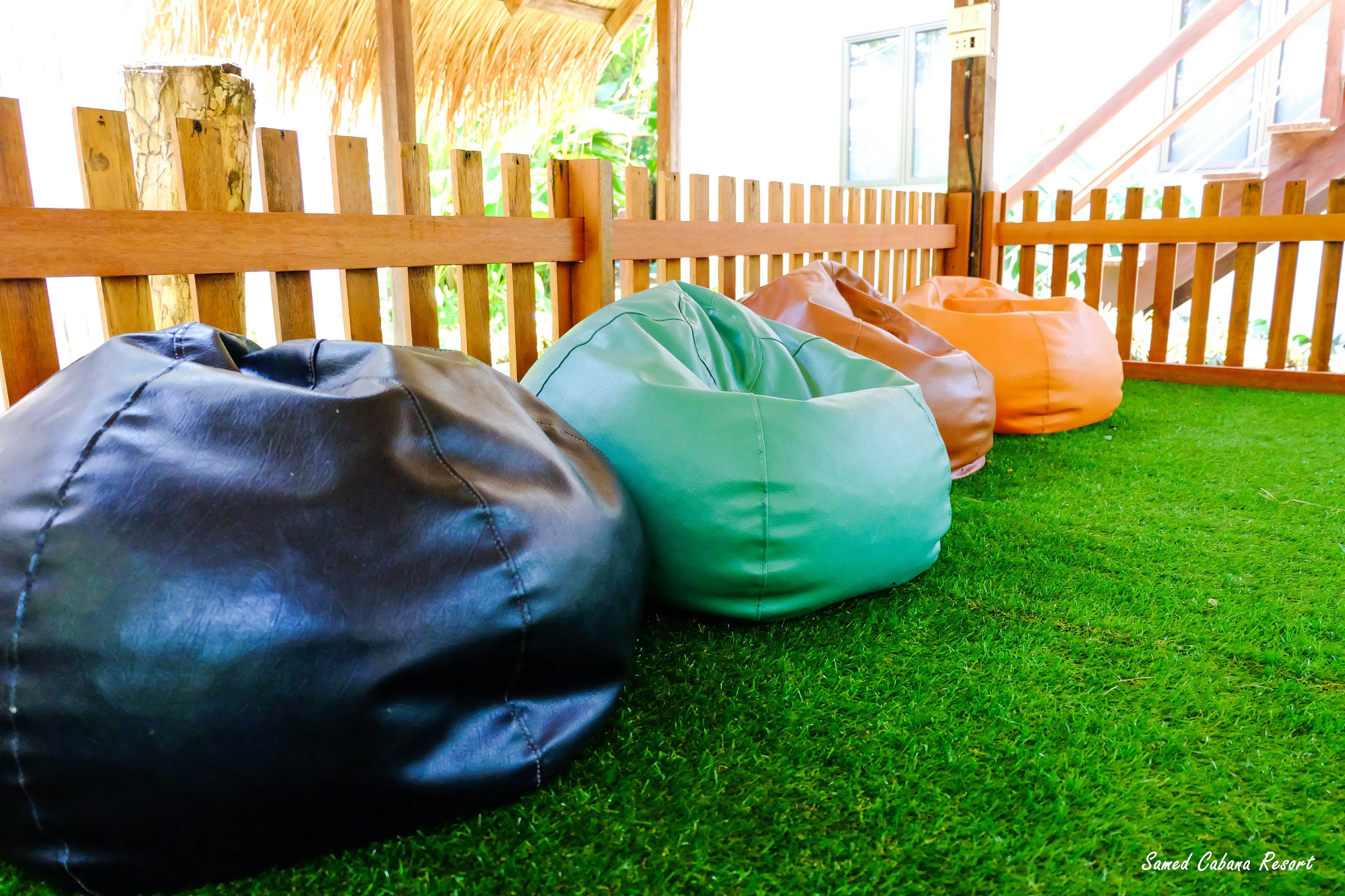Mobiliario exterior con sillones de mimbre sobre césped artificial, cerca de madera y estructura de techo de paja.