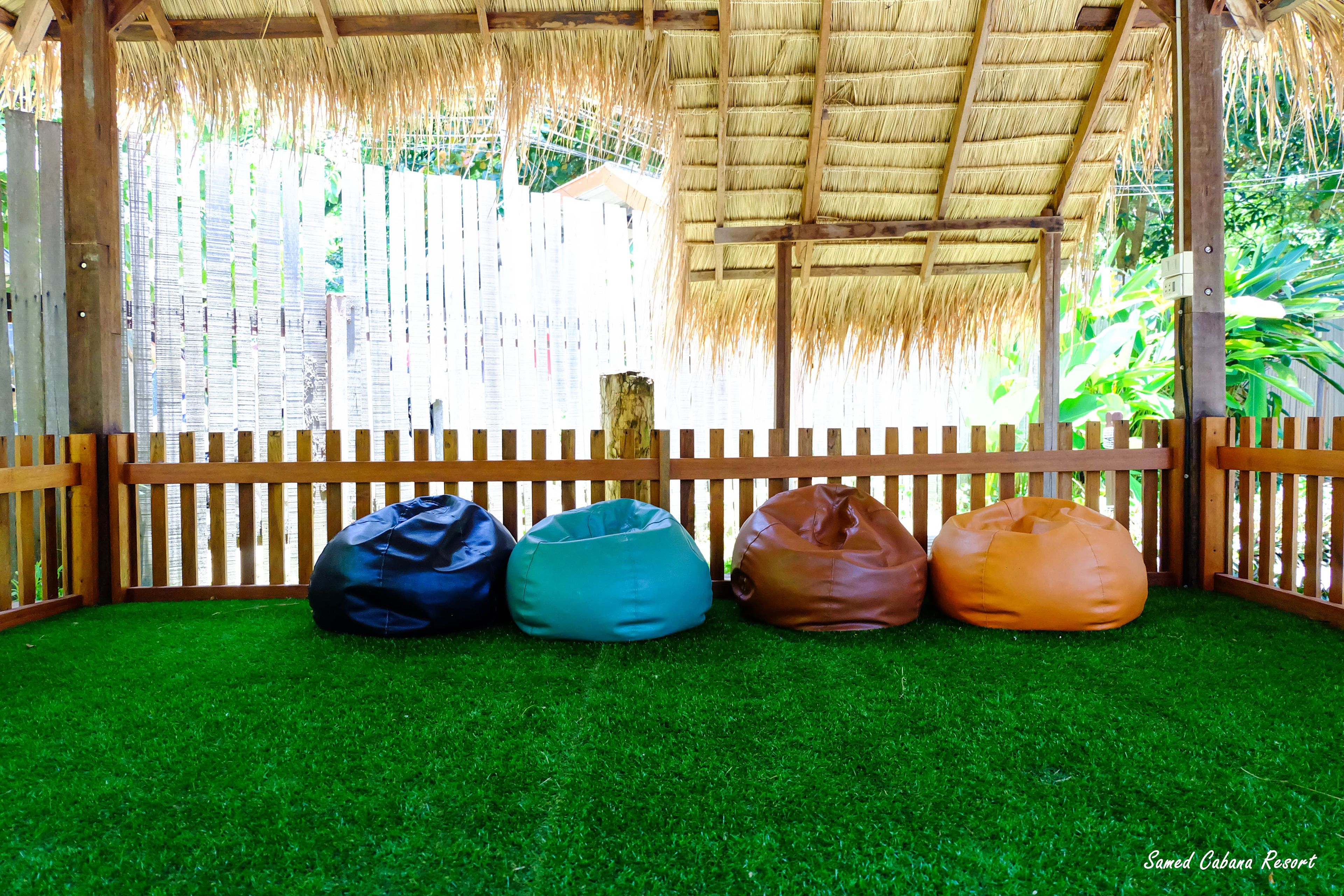A covered seating area with four bean bags on artificial grass, a wooden fence, and a thatched roof.