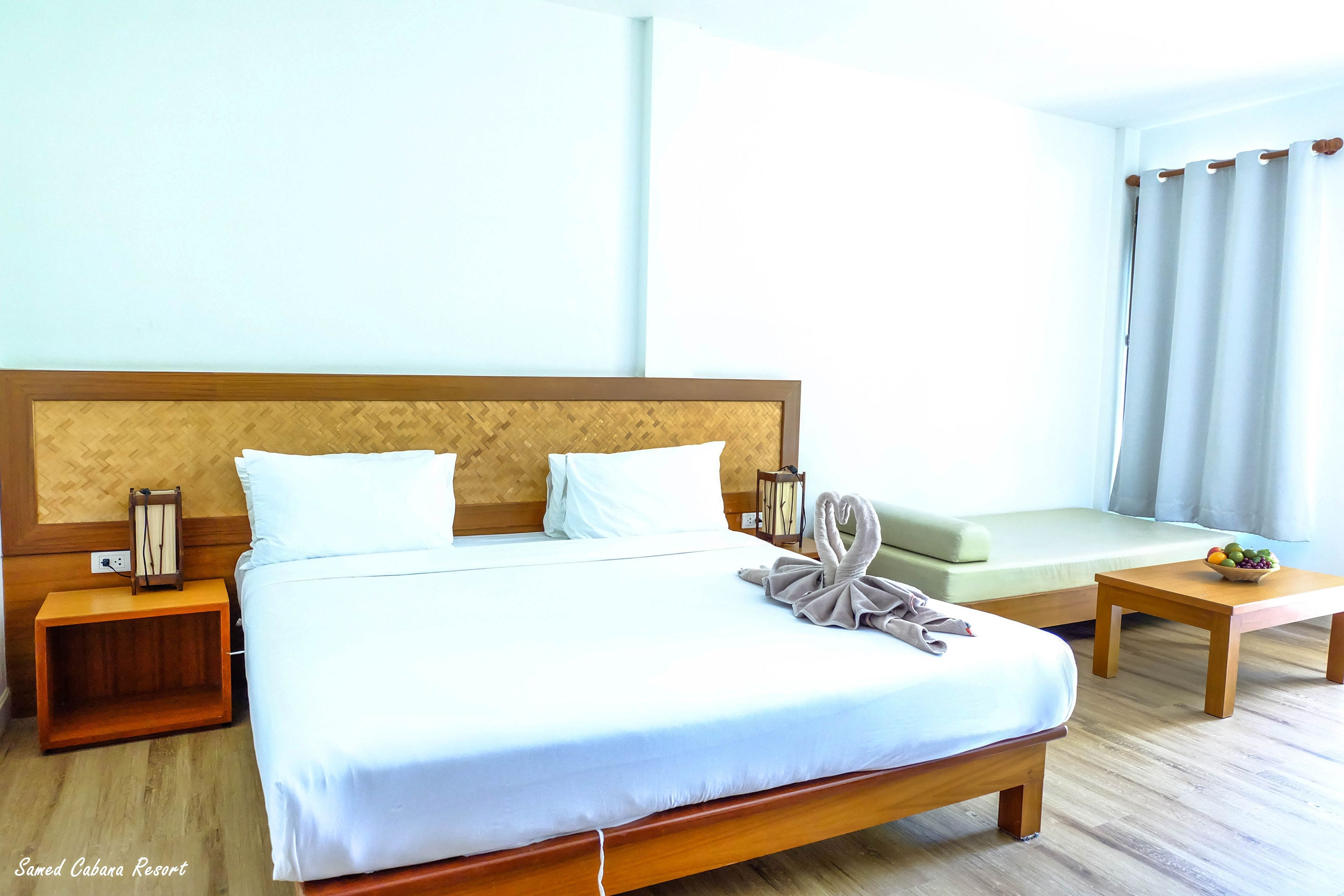 A hotel room with a large bed, a wooden headboard, a bedside table with a lamp, a small table with fruit, and a sofa.