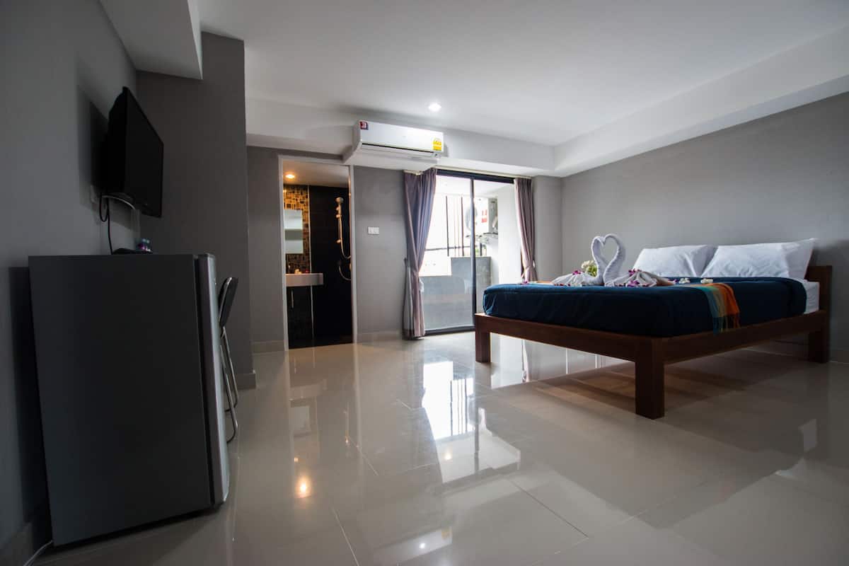 A modern hotel room with a bed, a flat-screen TV, a refrigerator, and a sliding glass door leading to a balcony.