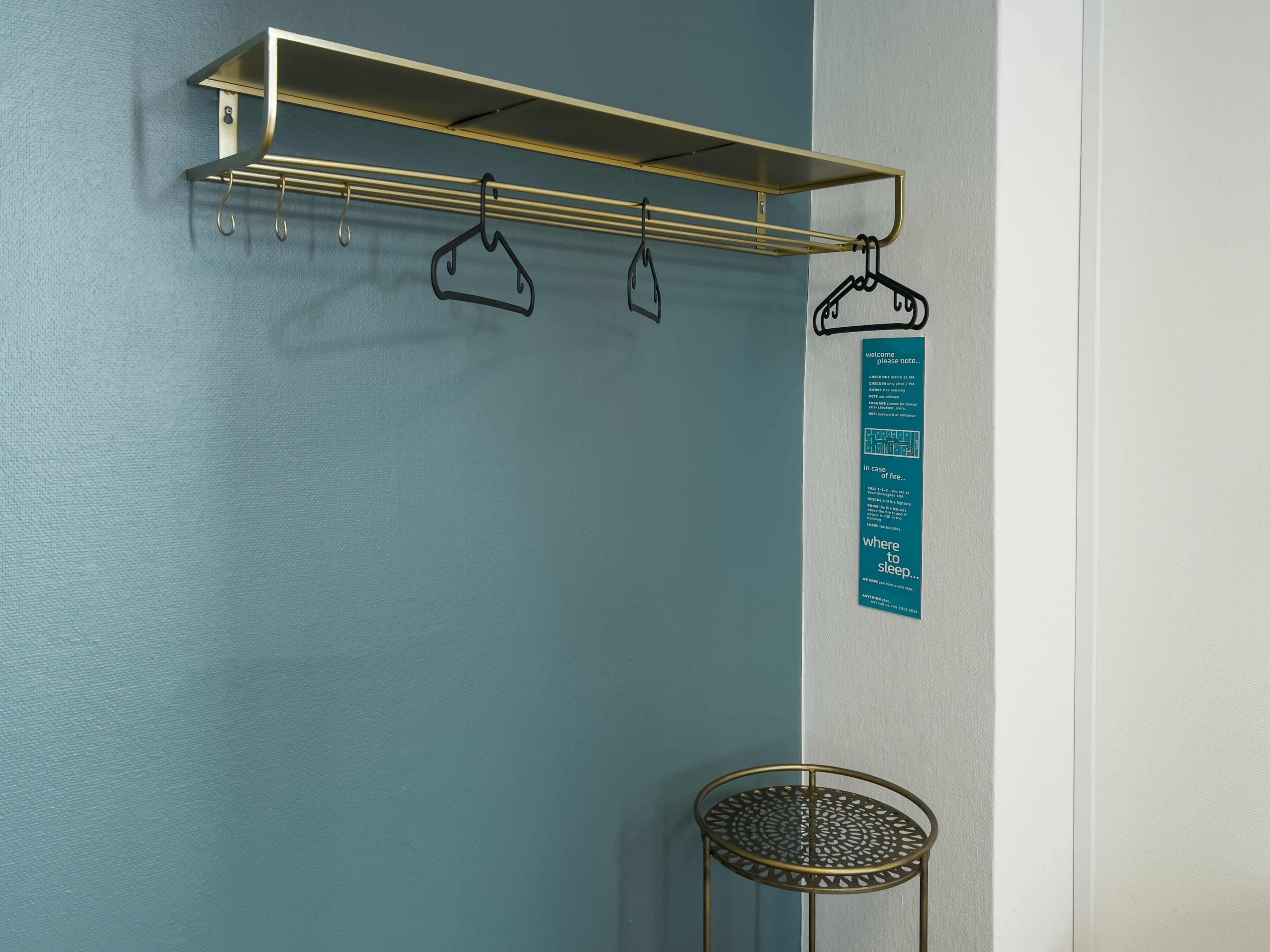 Een kamer met een blauwgroene muur, een gouden metalen plank met kledinghangers en een blauw informatieteken.