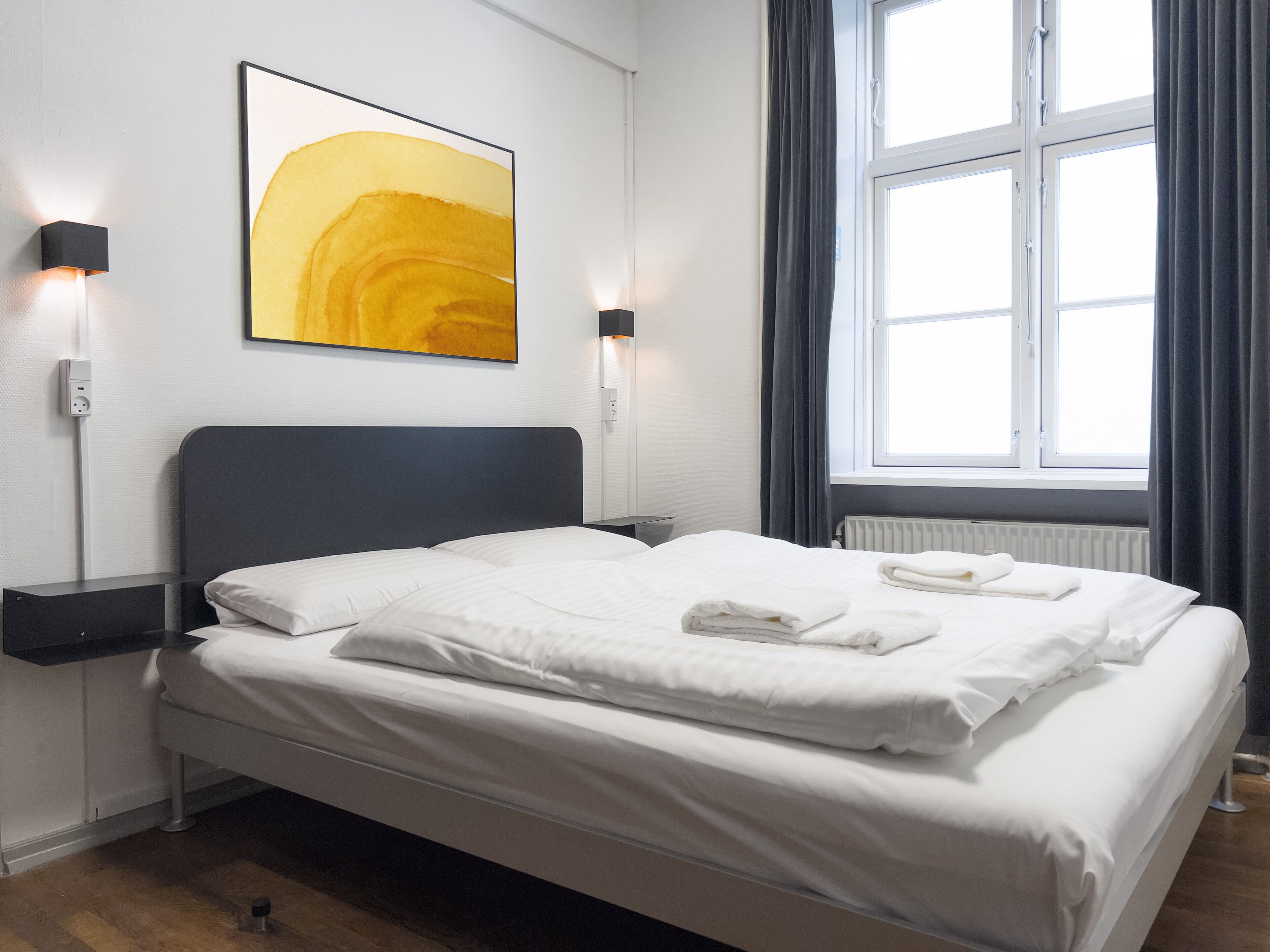 Una cama bien hecha con ropa blanca y toallas, un cabecero moderno y un amplio ventanal con cortinas.