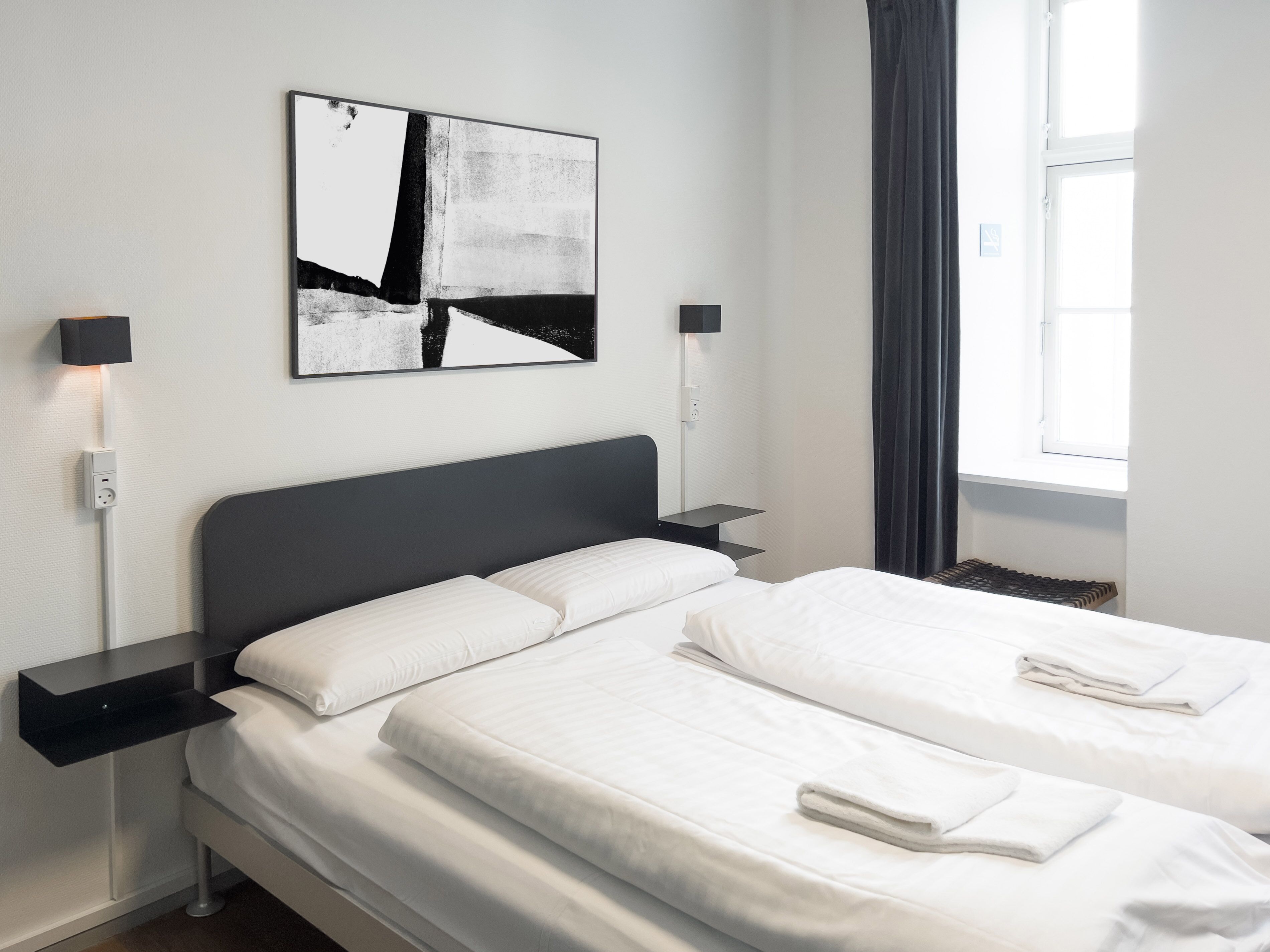 A modern hotel room with a large bed, bedside tables, a wall-mounted abstract painting, and a window with curtains.