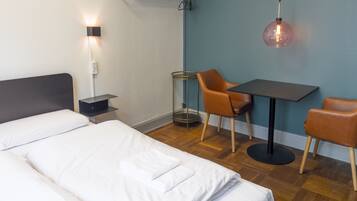A hotel room with a bed, a desk, two chairs, a lamp, and a wall-mounted shelf.