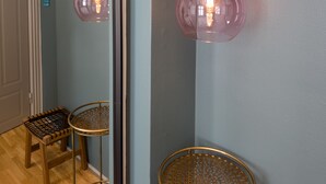 Esquina de una habitación con pared azul, suelo de madera, una silla metálica redonda y dos lámparas colgantes de tono rosado.