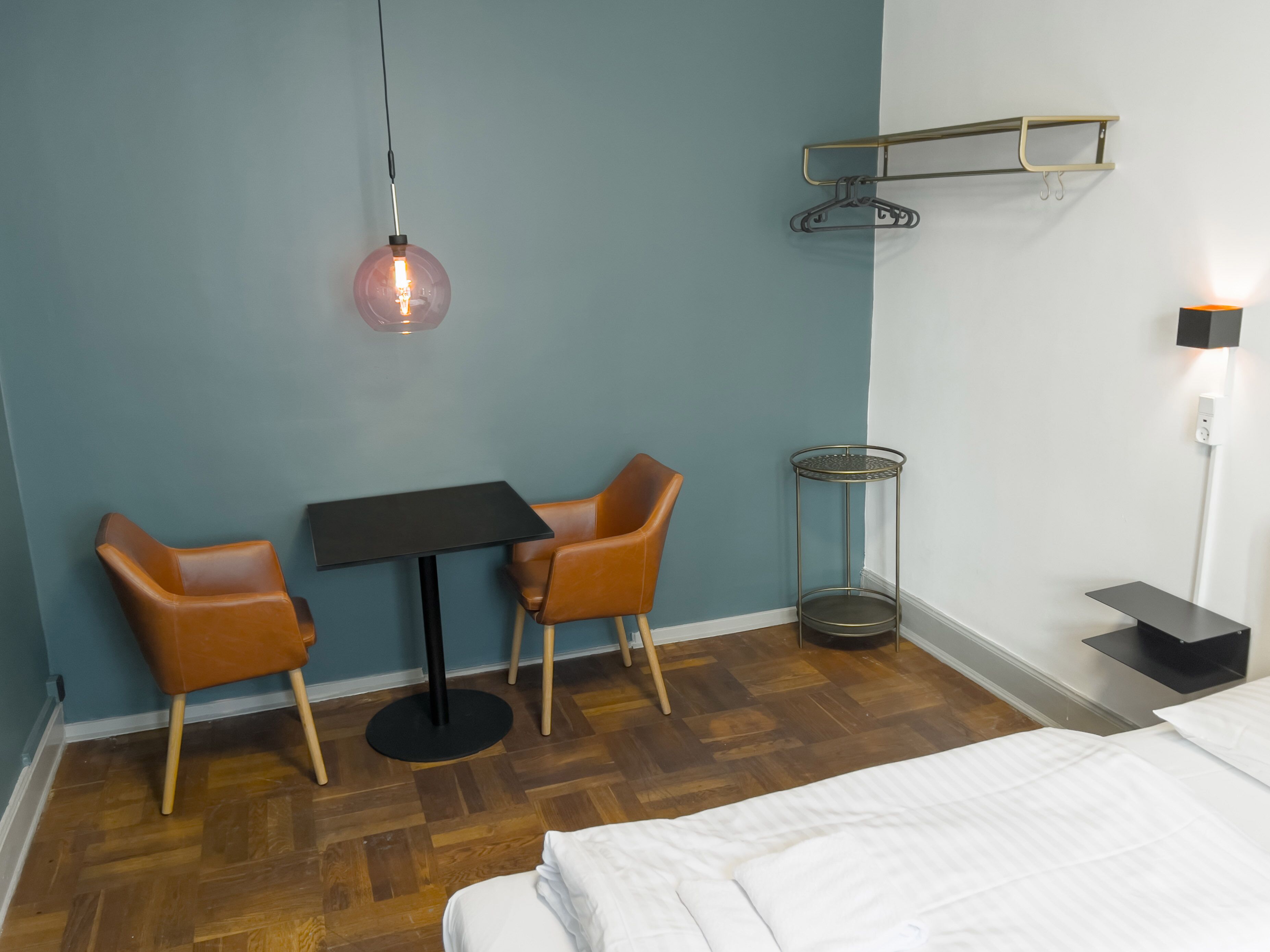 Habitación de hotel con cama, dos sillas, una mesita, una lámpara colgante, una mesita de noche y un estante en la pared.
