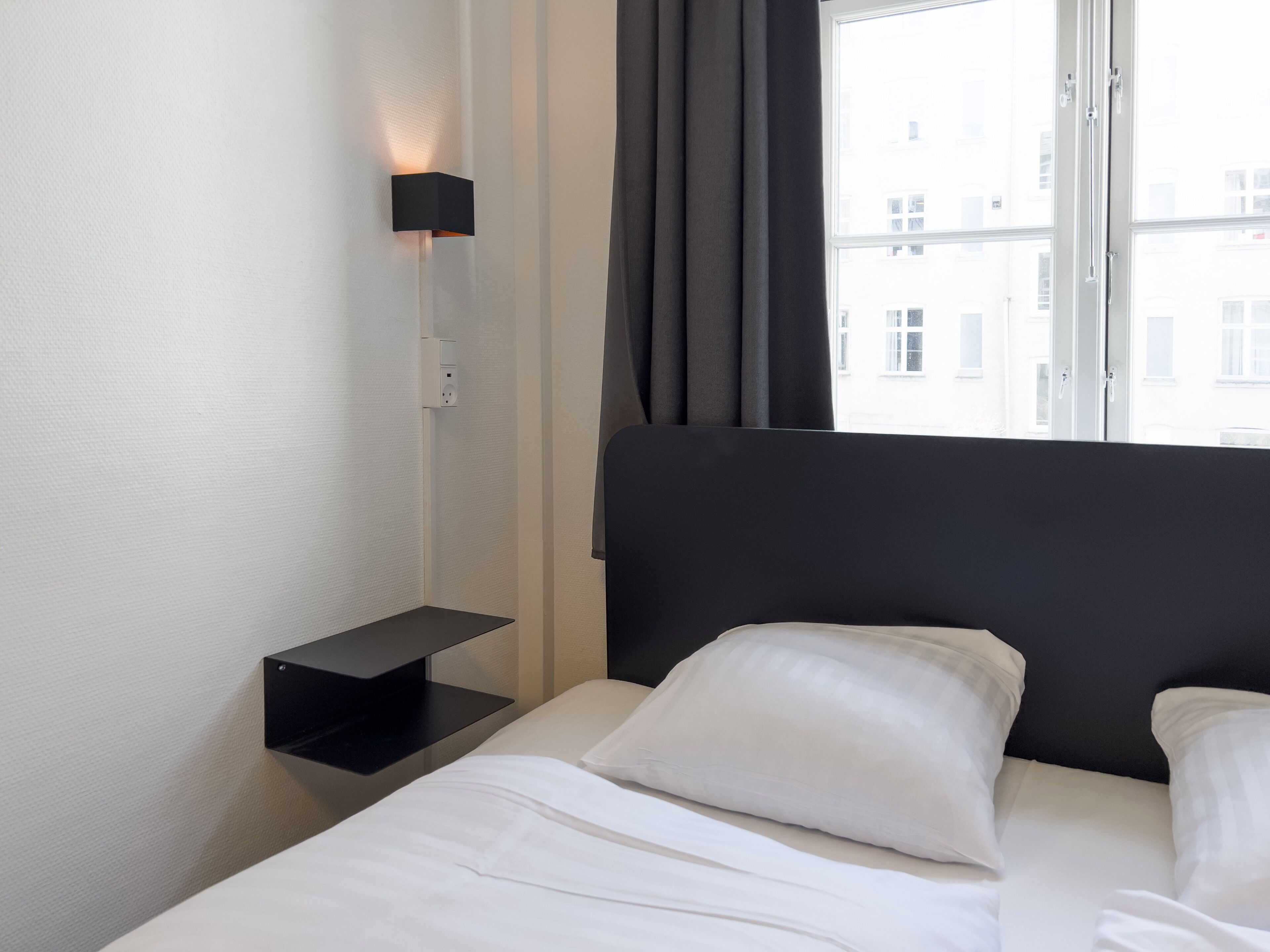 A hotel room with a bed, a bedside table, a wall-mounted light, and a window with curtains.