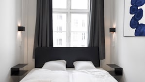 Habitación de hotel moderna con una cama grande, cabecera oscura y ropa de cama blanca. Hay dos mesitas de noche con lámparas, una ventana con cortinas y una obra de arte abstracta en la pared.