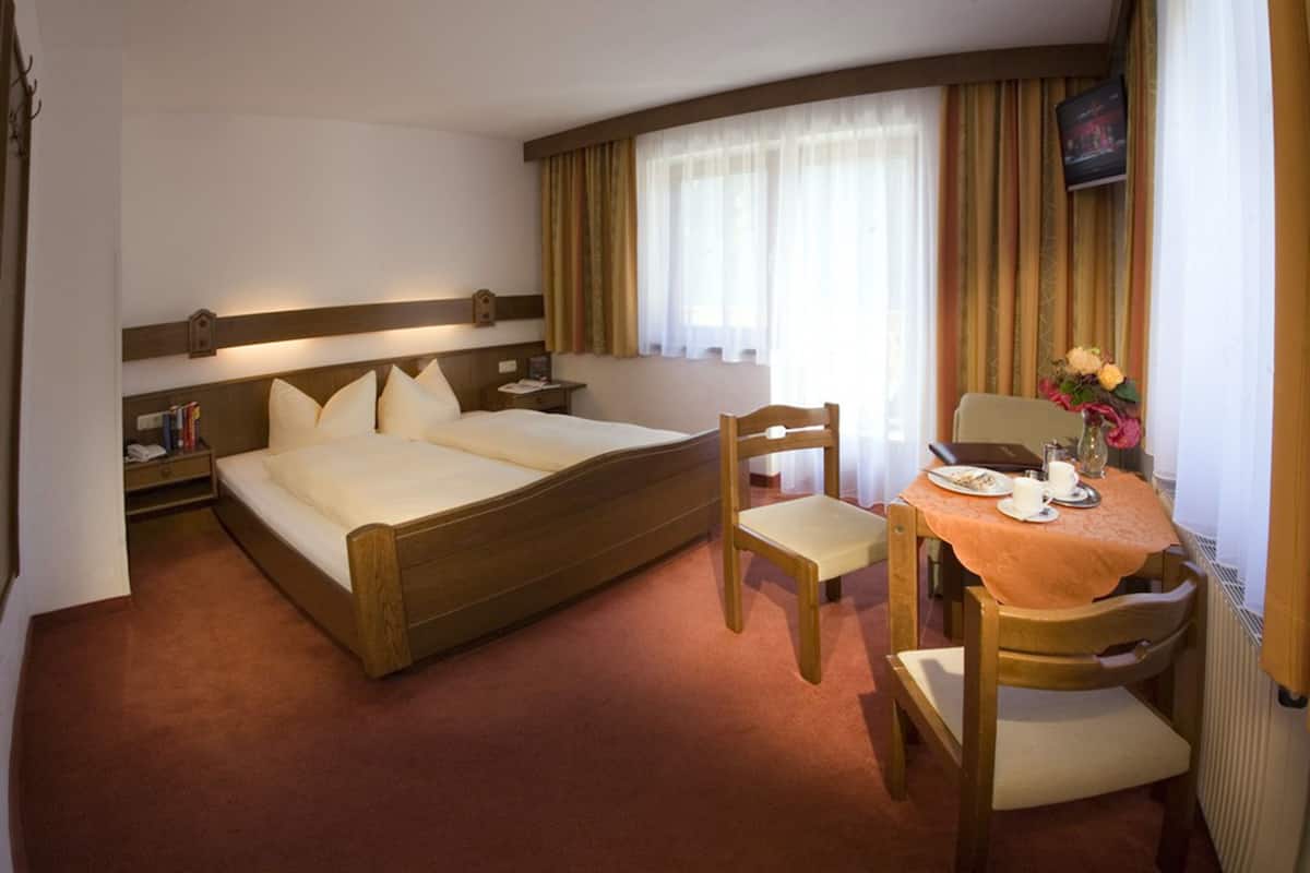 Camera d'albergo con un letto matrimoniale, una sedia in legno, un tavolino con un vaso di fiori e un televisore a parete.