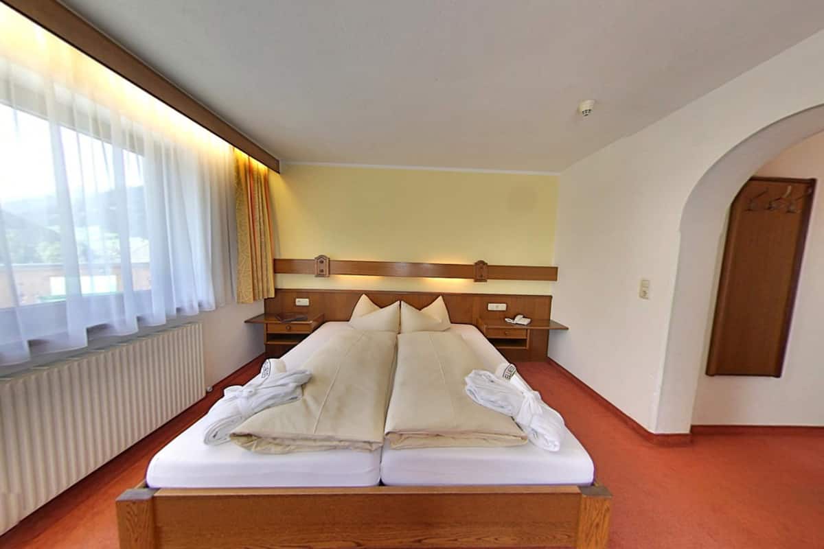 Un letto matrimoniale con lenzuola e asciugamani bianchi, una testiera in legno, un comodino con una lampada e una finestra con tende leggere.