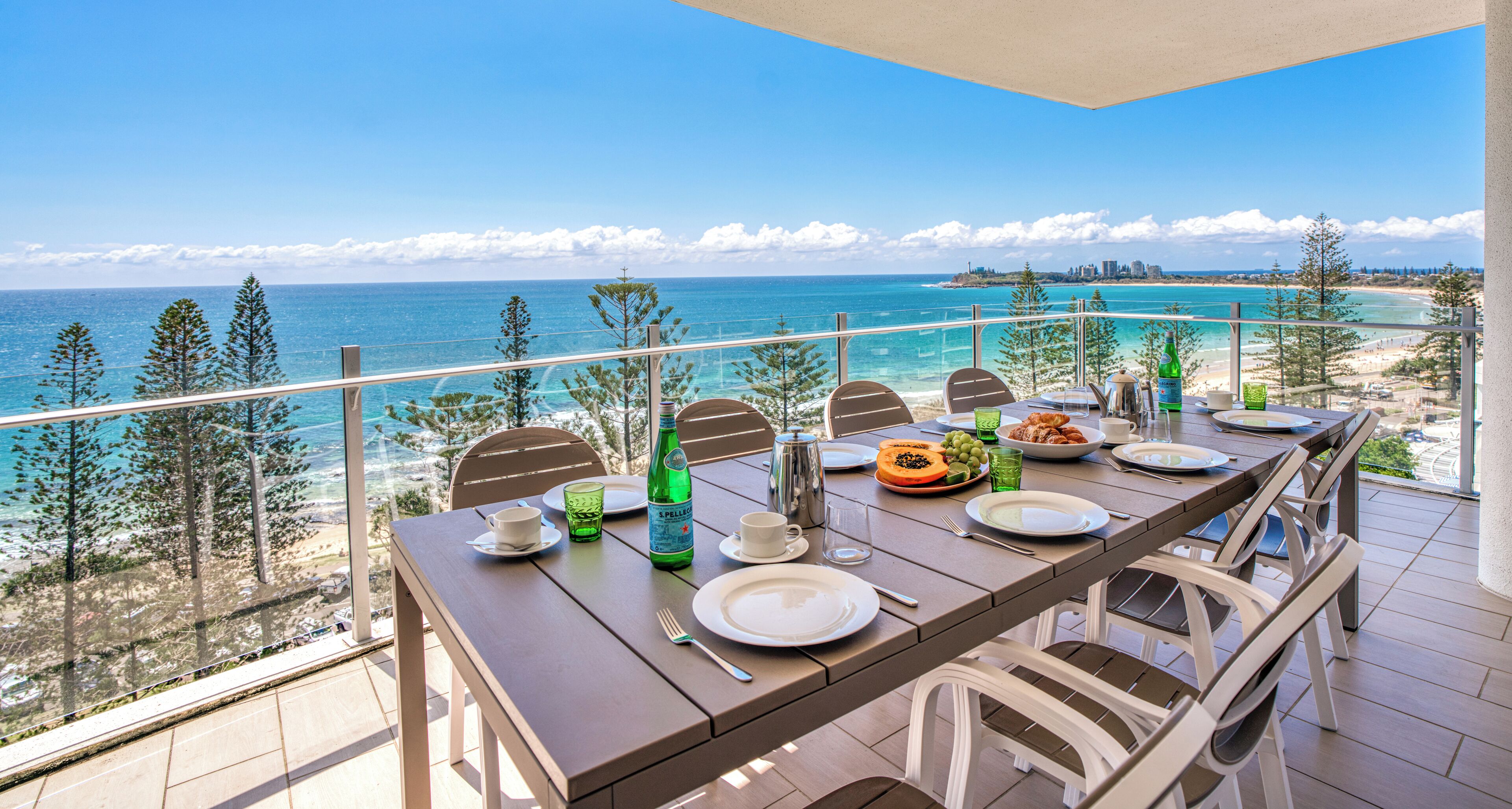 A dining table set on a balcony with a view of the ocean and beach.