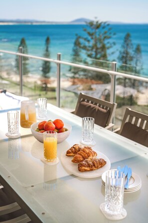 A breakfast table with croissants, a bowl of fruit, and a glass of orange juice, overlooking a scenic ocean view.