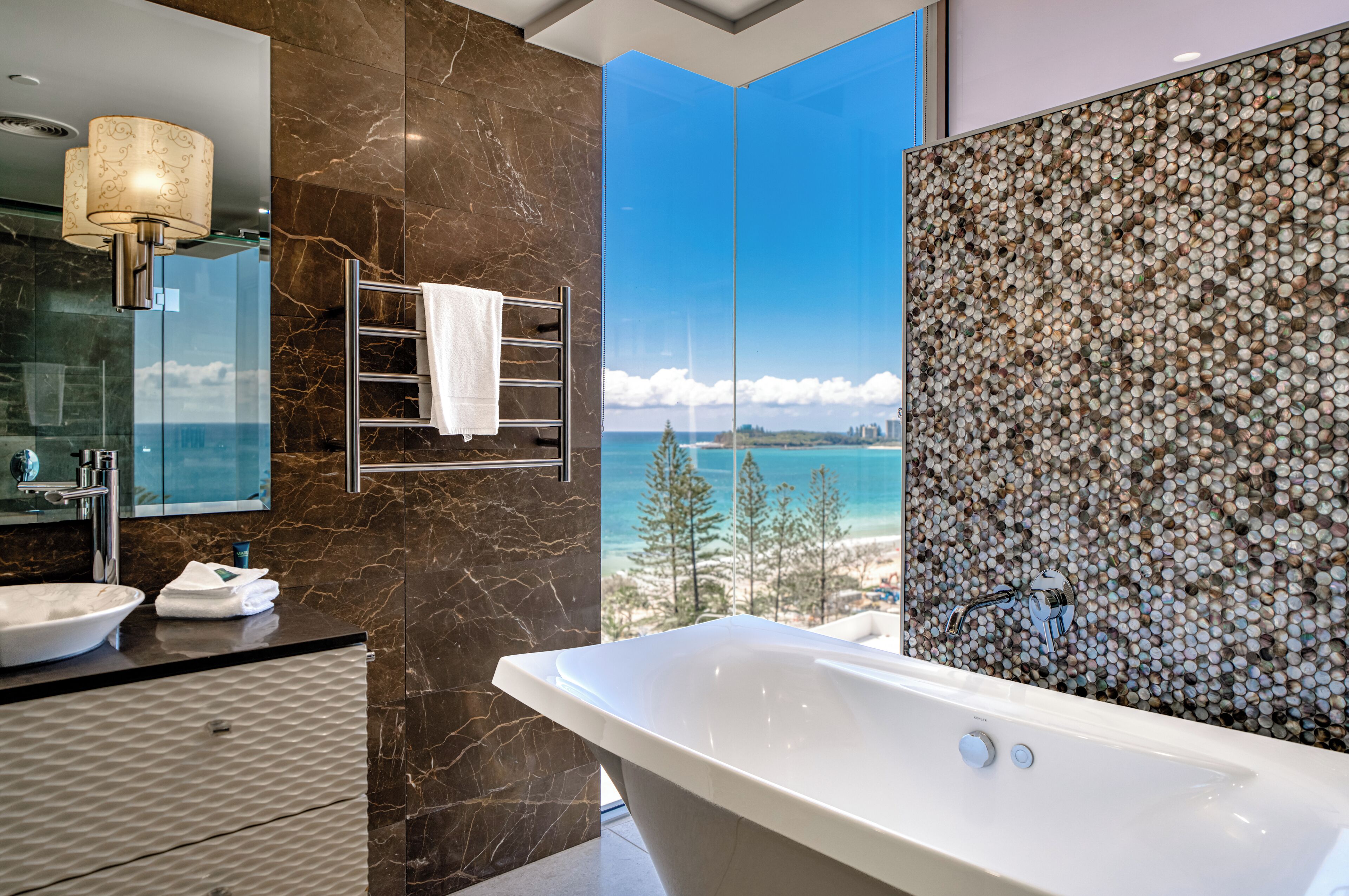 A modern bathroom with a large bathtub, a sink, and a mirror. There is a view of the beach and ocean through a glass partition.