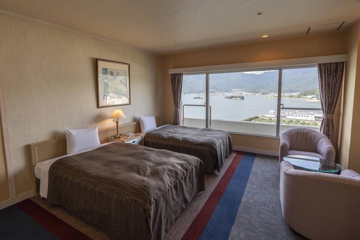 Een hotelkamer met twee bedden, een stoel, een tafel, een lamp en een groot raam met uitzicht op een waterpartij en bergen.