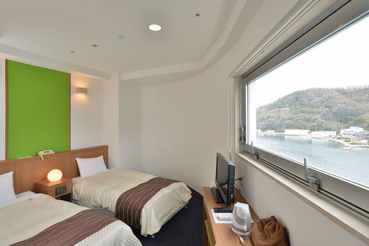 Een hotelkamer met twee bedden, een bureau met een computer, en een raam met uitzicht op een waterpartij en heuvels.
