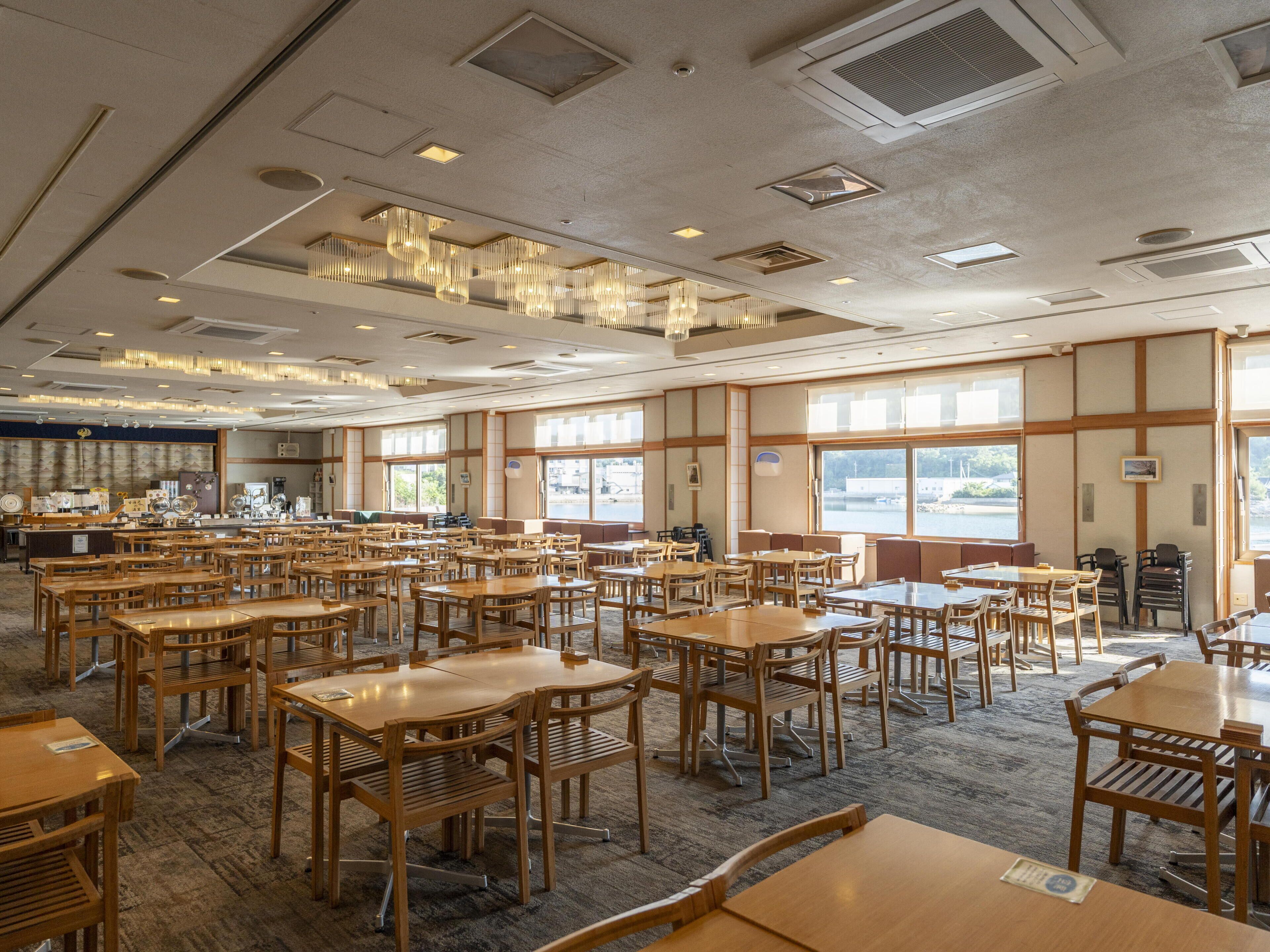 A spacious dining area with multiple wooden tables and chairs, large windows offering views, and a ceiling with recessed lighting and air vents.