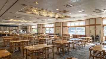 A spacious dining area with multiple wooden tables and chairs, large windows offering views, and a ceiling with recessed lighting and air vents.