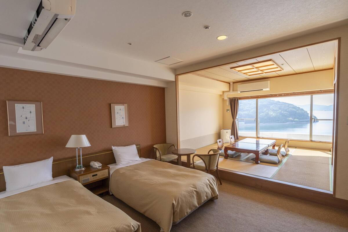 Hotelkamer met twee bedden, een bureau, een stoel en een groot raam met uitzicht op een wateroppervlak en een berg.