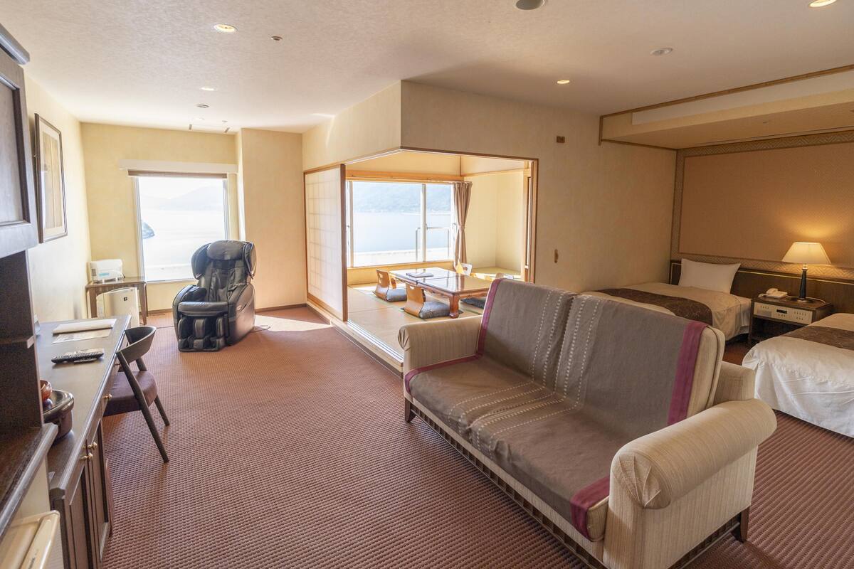 Een hotelkamer met een bank, twee bedden, een massagestoel, een bureau met een computer en uitzicht op zee.