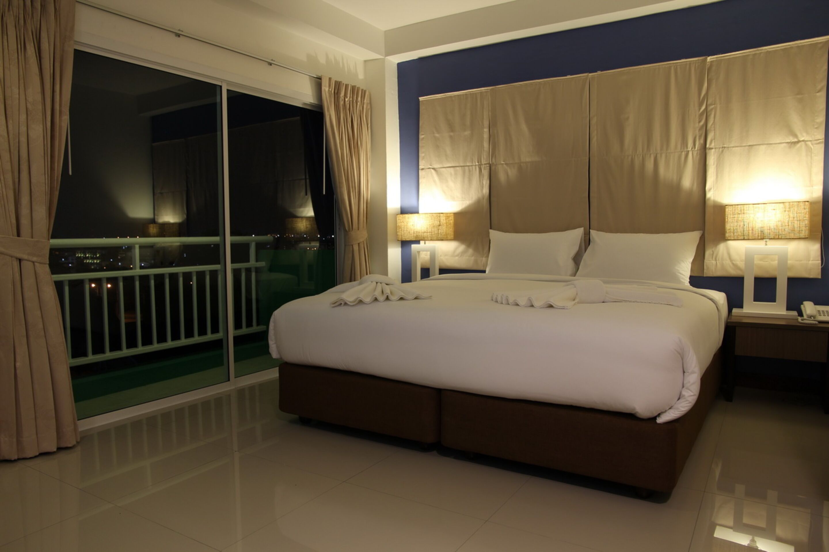 A hotel room with a large bed, two bedside tables, a nightstand, a balcony with curtains, and a city view.