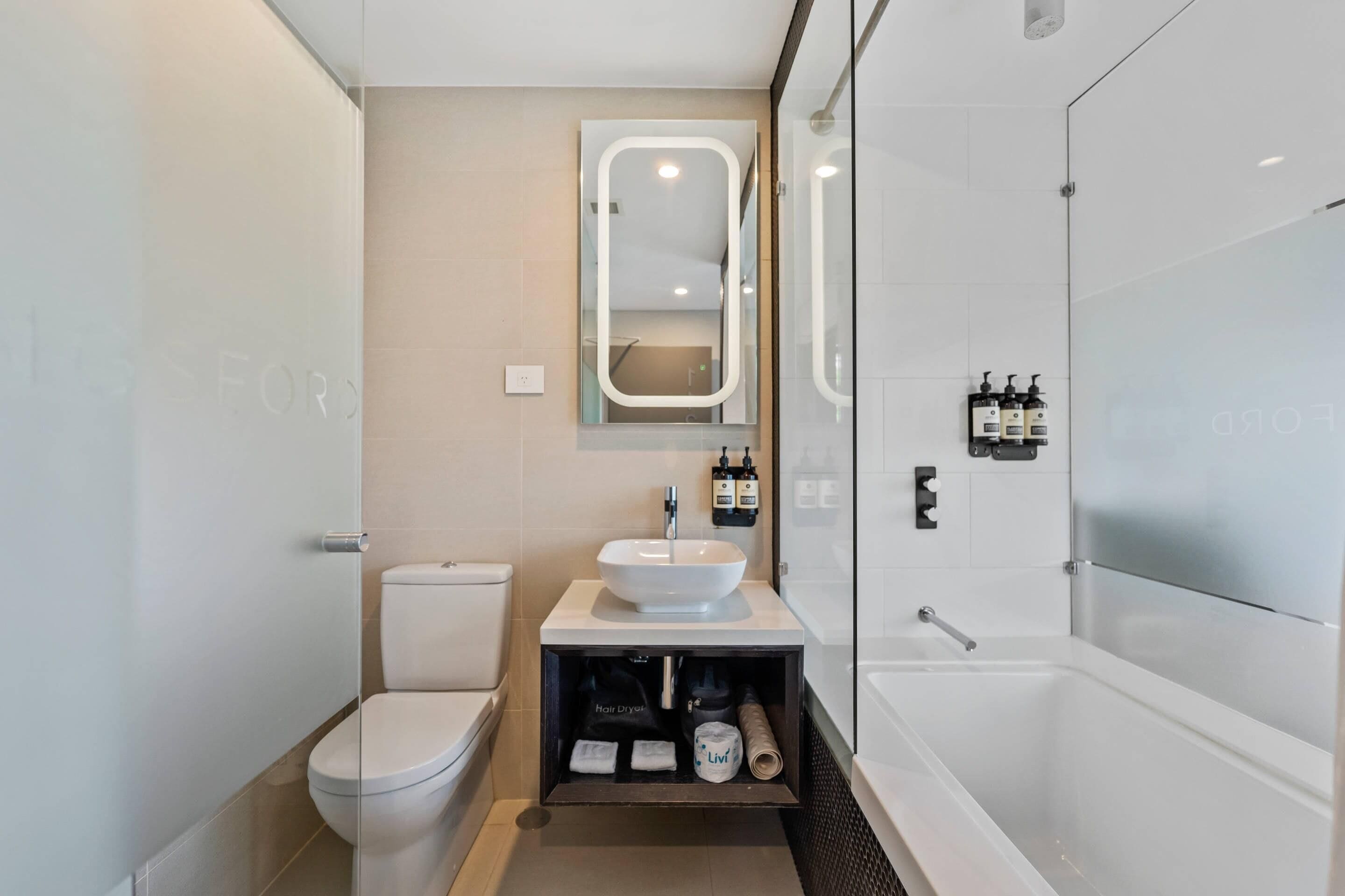 A modern bathroom with a toilet, sink, and bathtub. There is a mirror above the sink and a shelf with toiletries.