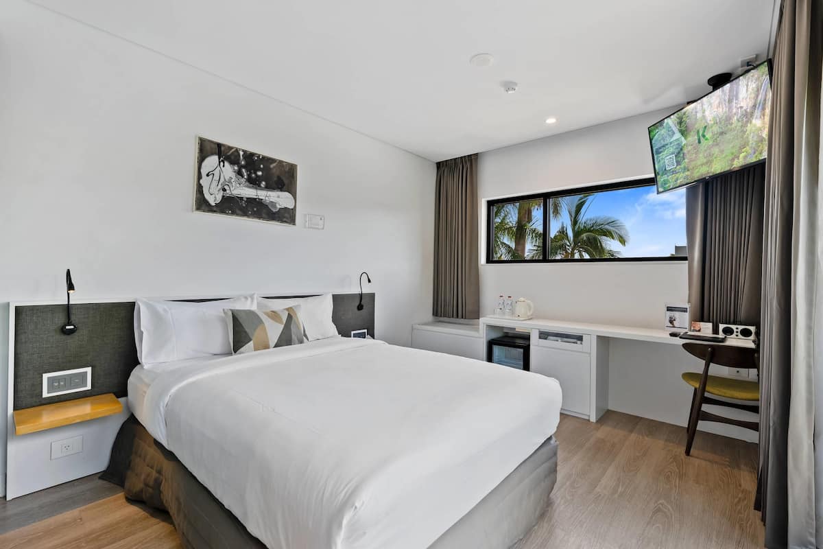 Habitación de hotel moderna con una cama grande, un escritorio con computadora, un televisor y una ventana con vistas a palmeras.