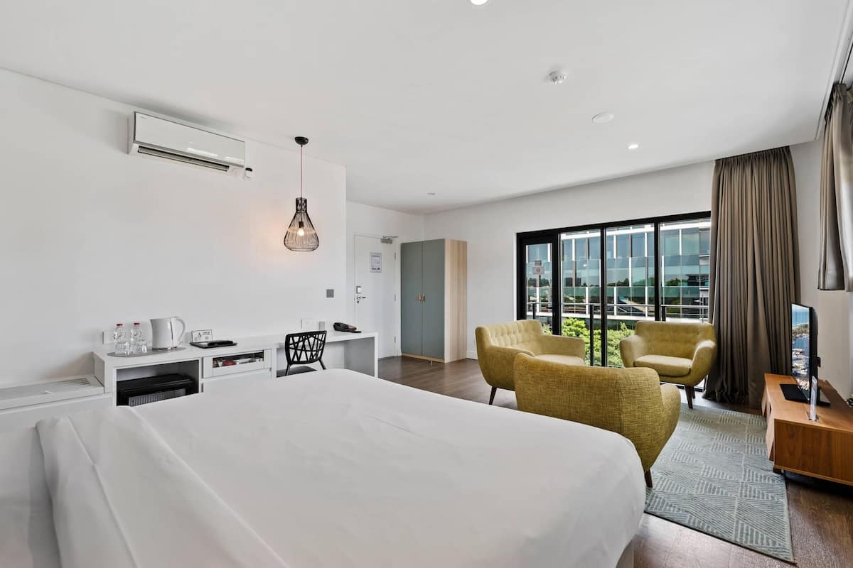 Habitación de hotel moderna con una cama grande, dos sillones, un escritorio con computadora y un ventanal con vistas a la ciudad.