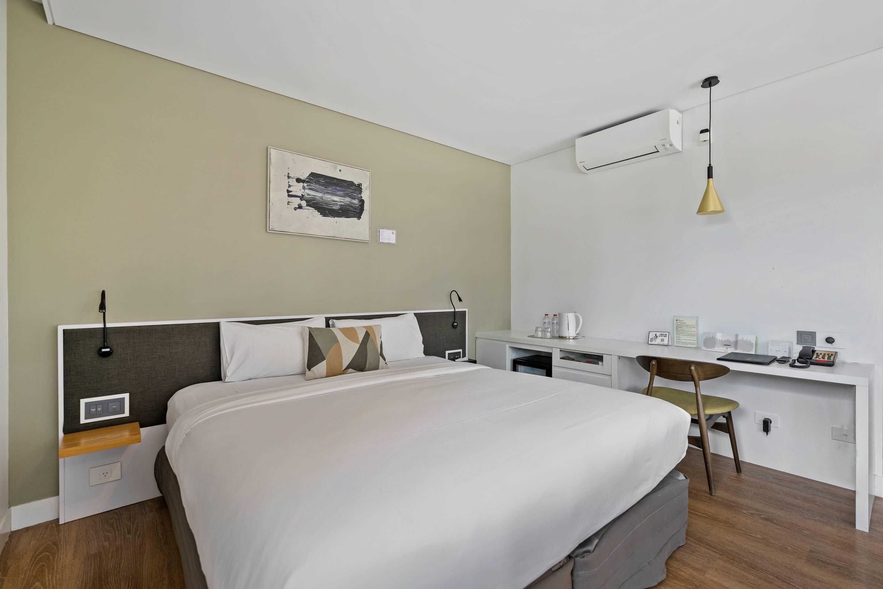 A hotel room with a bed, bedside table, desk, chair, and a wall-mounted air conditioner.