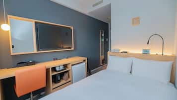 Ett hotellrum med en säng, ett skrivbord med en stol, ett litet kylskåp, en spegel, ett nattduksbord med en lampa och en väggmonterad TV.