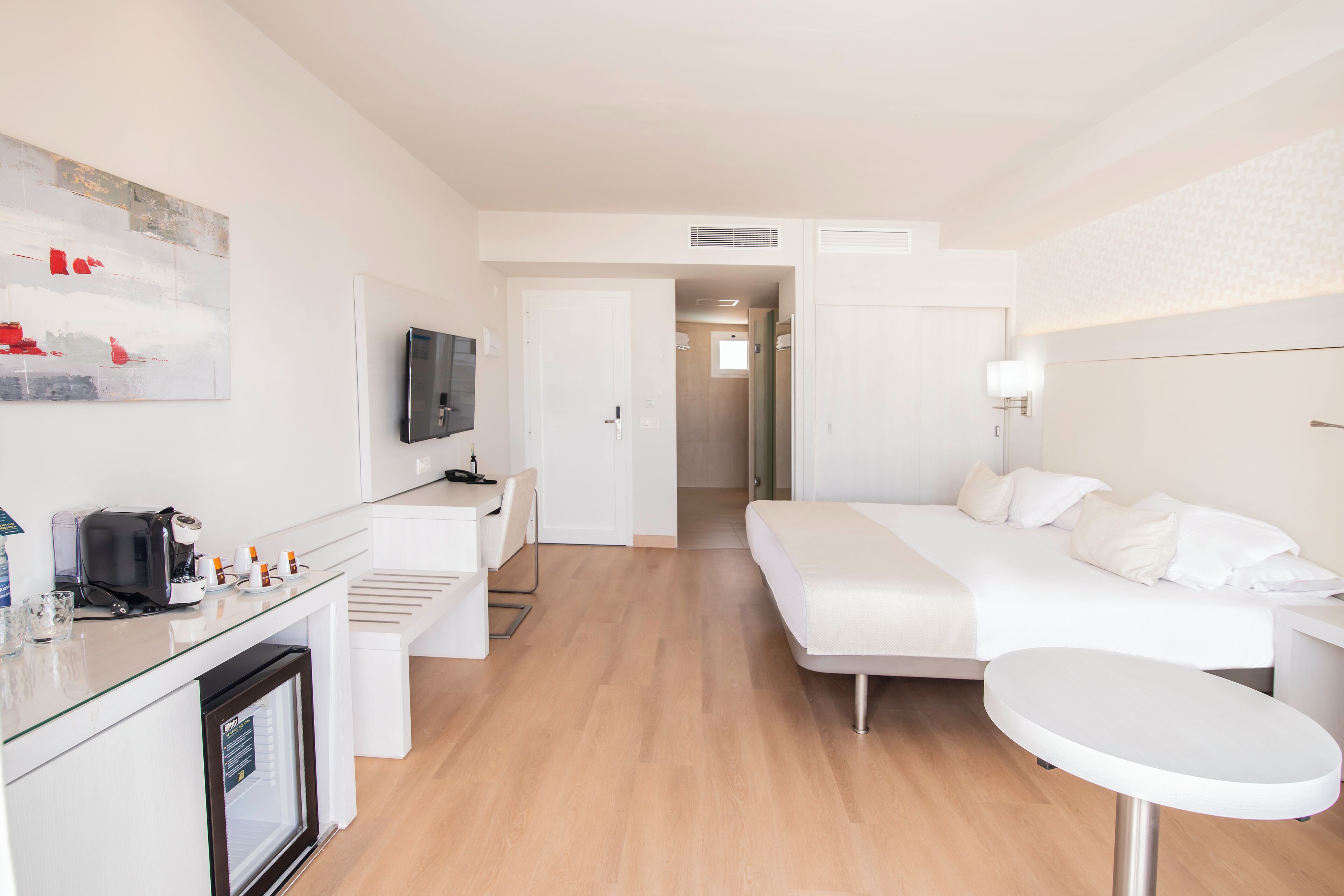 A modern hotel room with a bed, a desk with a computer, a coffee machine, and a TV.