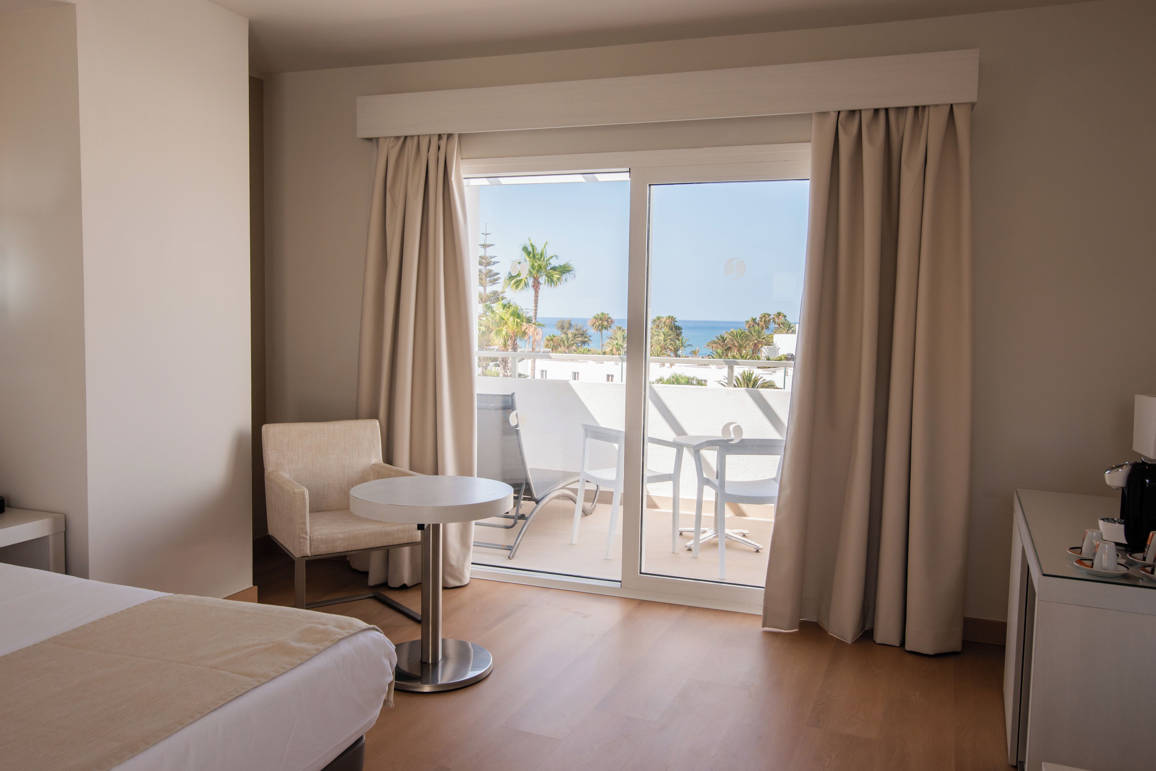 Habitación de hotel con cama, una mesita, una silla y un balcón con vistas a palmeras y al mar.