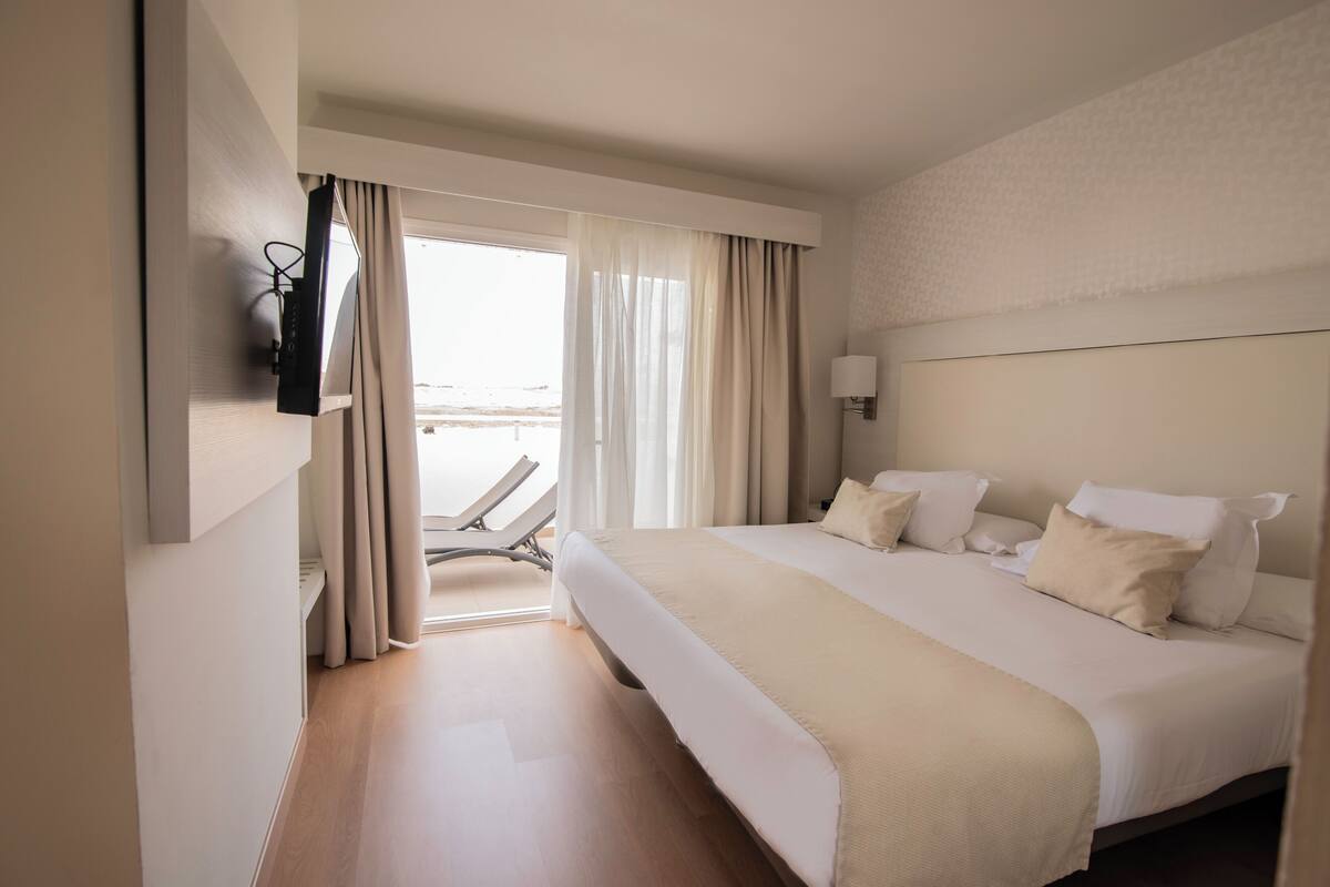 Habitación de hotel con una cama grande, un televisor montado en la pared y vista al mar por la ventana.