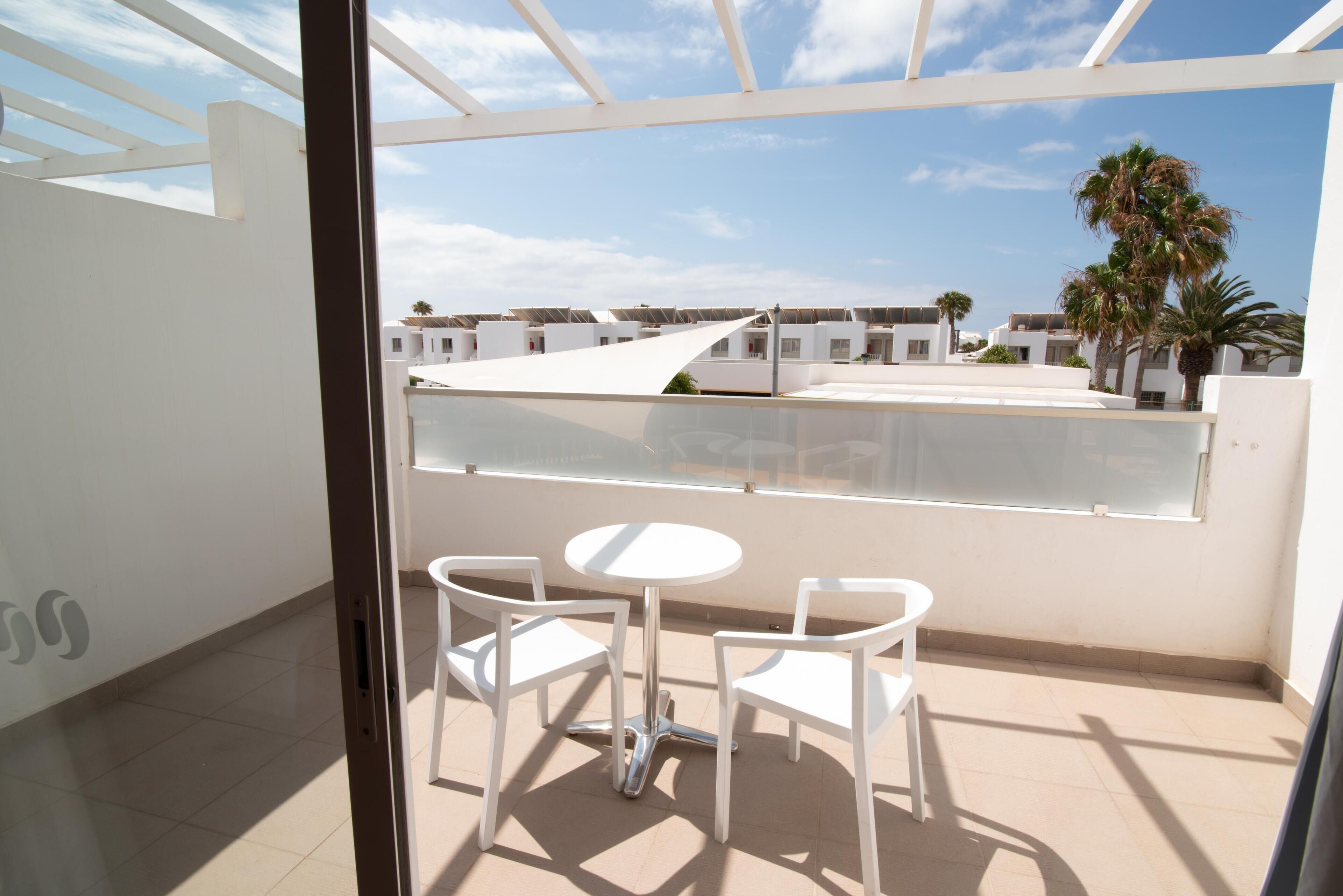 Un balcon avec une table et des chaises blanches, une vue sur des immeubles et des palmiers, et un ciel dégagé.