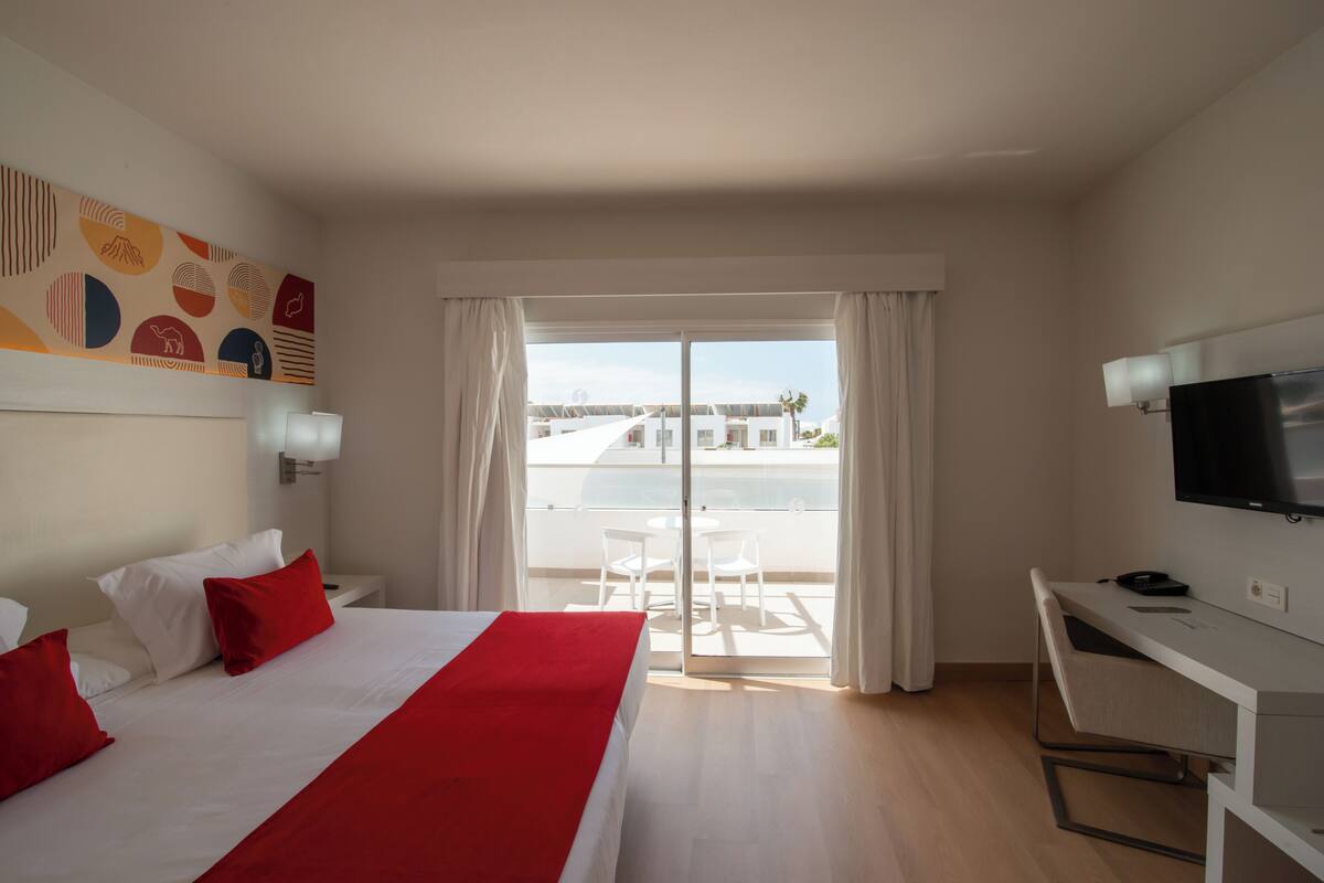 Habitación de hotel con cama, escritorio, televisión y balcón con vistas.