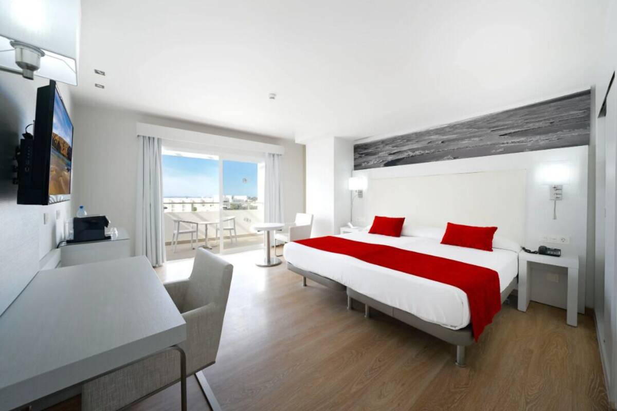 Habitación de hotel con cama, escritorio, silla, mesita pequeña, mesita de noche y vista al exterior.