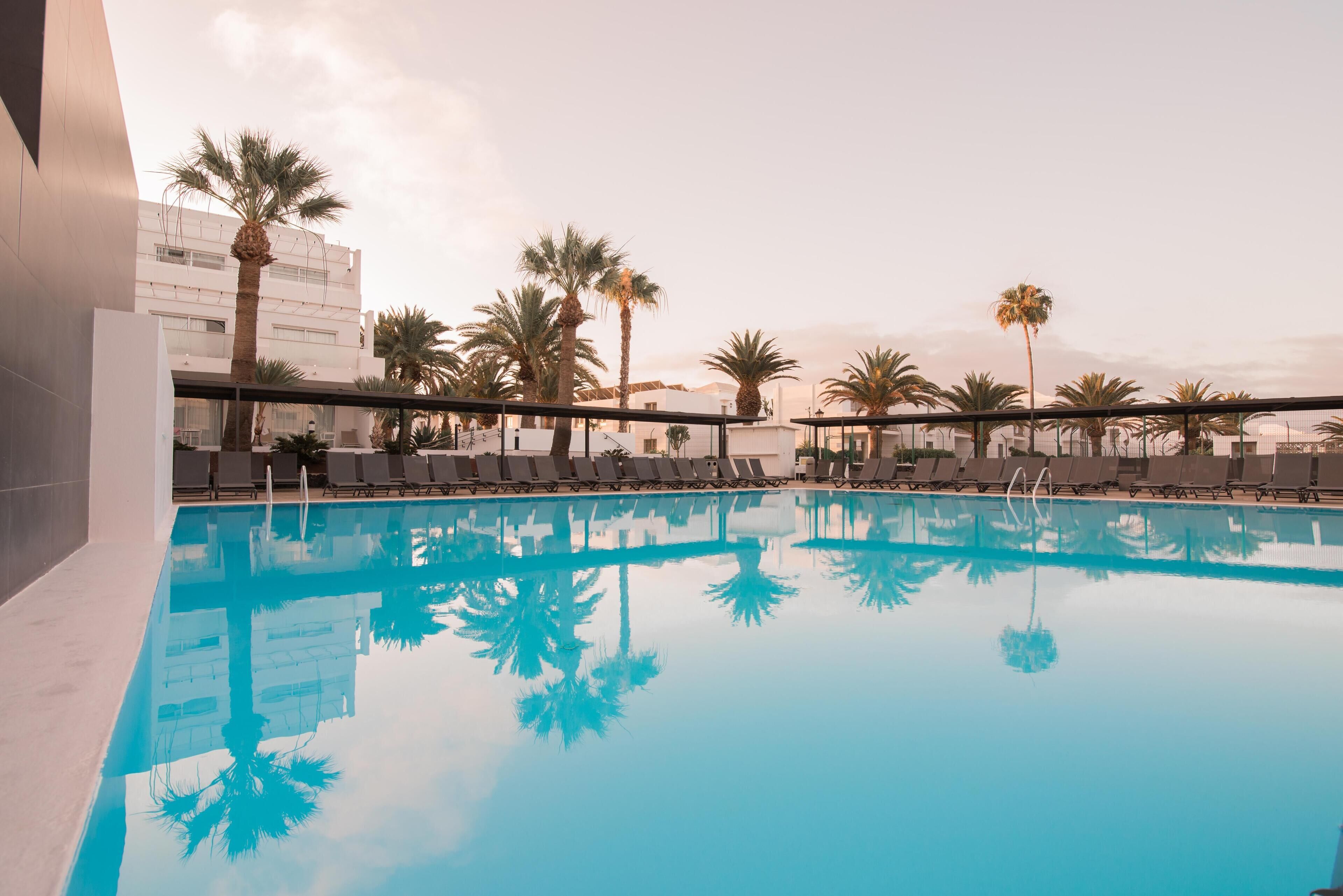 Una piscina con sillones de descanso y palmeras, rodeada de edificios.