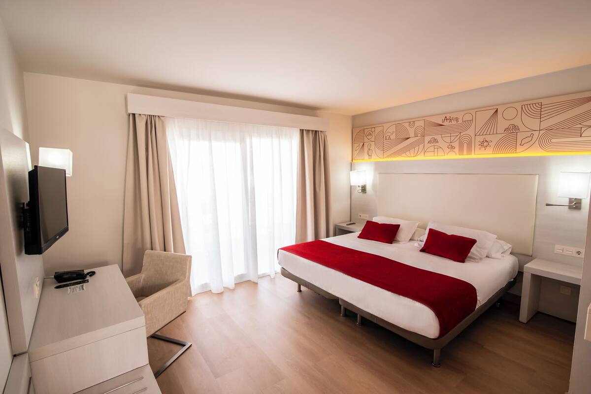 Habitación de hotel con una cama grande, un escritorio, una silla, un televisor y una ventana con cortinas.