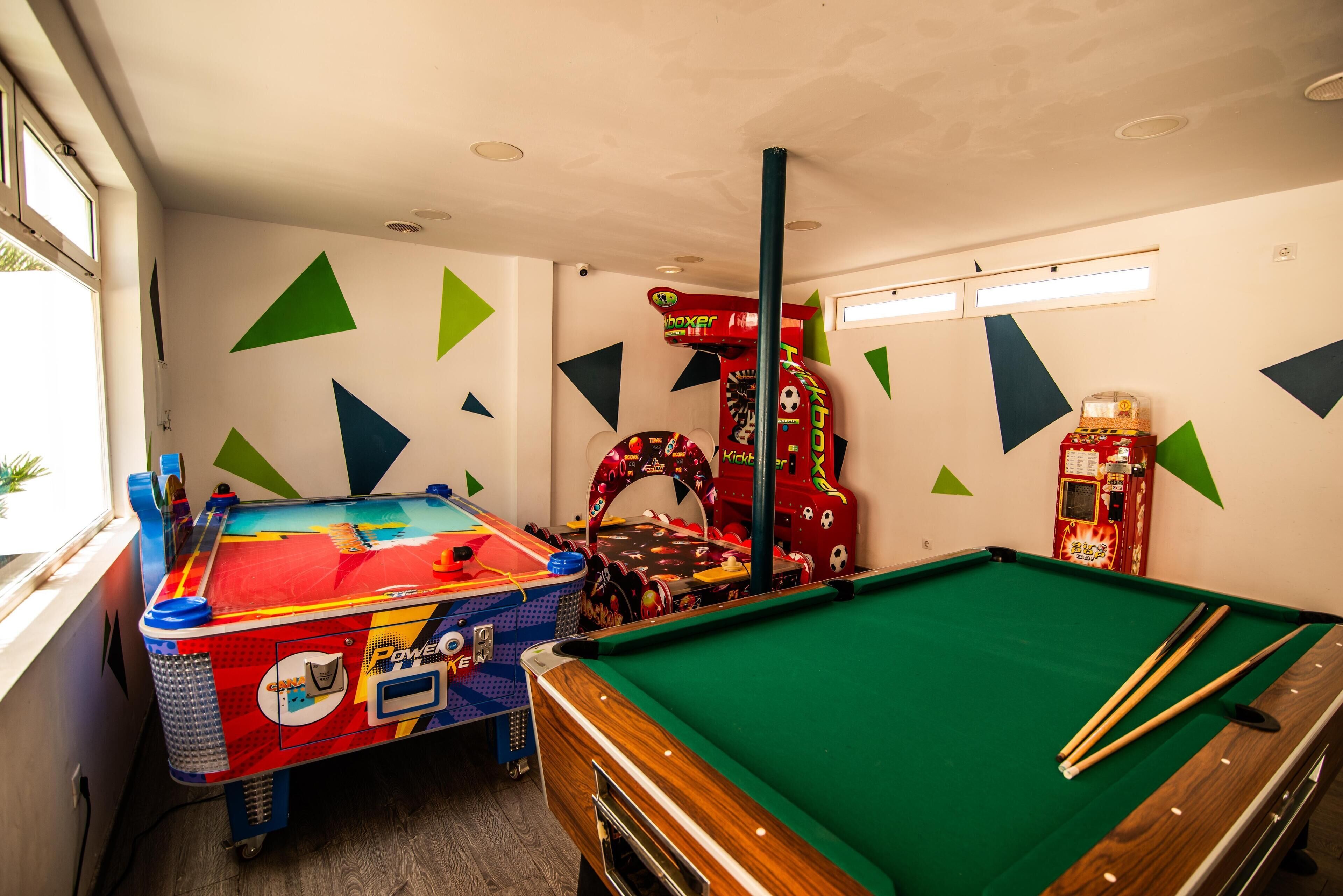 Una mesa de billar con superficie de fieltro verde, una mesa de pool colorida con diseño azul y rojo, y una máquina arcade roja.