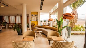 A modern hotel lobby with a curved sofa, comfortable seating, a reception desk, and a large window offering a view of the outdoors. - Aequora Lanzarote Suites (Tías)