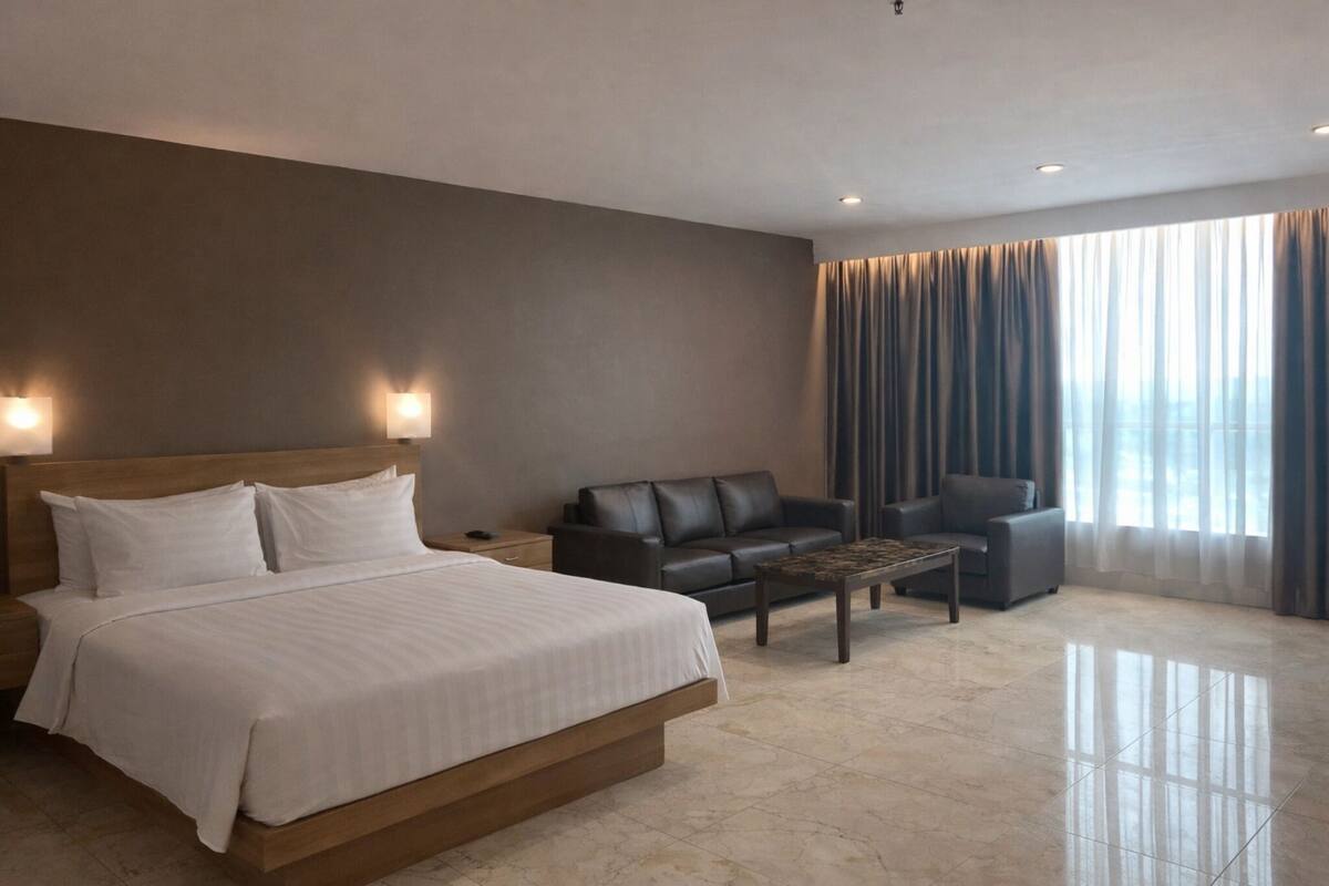 A hotel room with a large bed, a dark leather sofa, a coffee table, and a view of the city through sheer curtains.