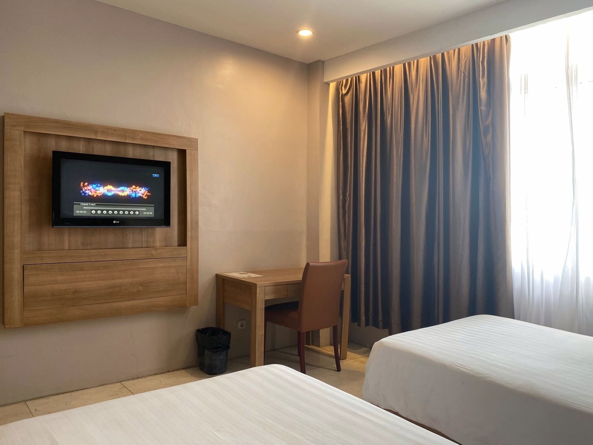 Habitación de hotel con una televisión montada en la pared, un escritorio, dos camas y una ventana con cortinas.