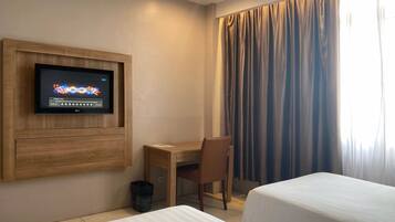 Habitación de hotel con una televisión montada en la pared, un escritorio, dos camas y una ventana con cortinas.