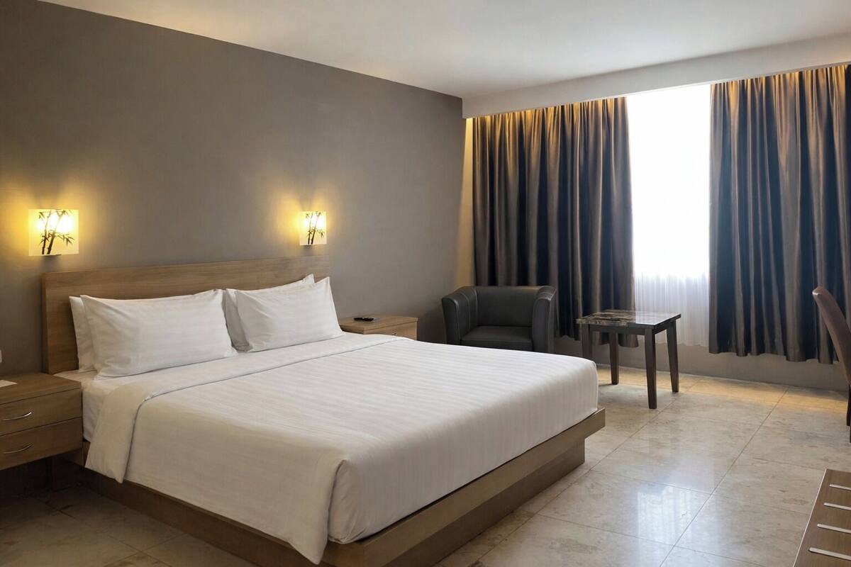 A hotel room with a large bed, two bedside tables, a chair, a small table, and a window with curtains.