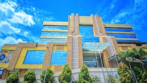 Un bâtiment moderne doté d’une façade en verre, une entrée centrale ornée de colonnes décoratives, et un ciel bleu clair en arrière-plan.