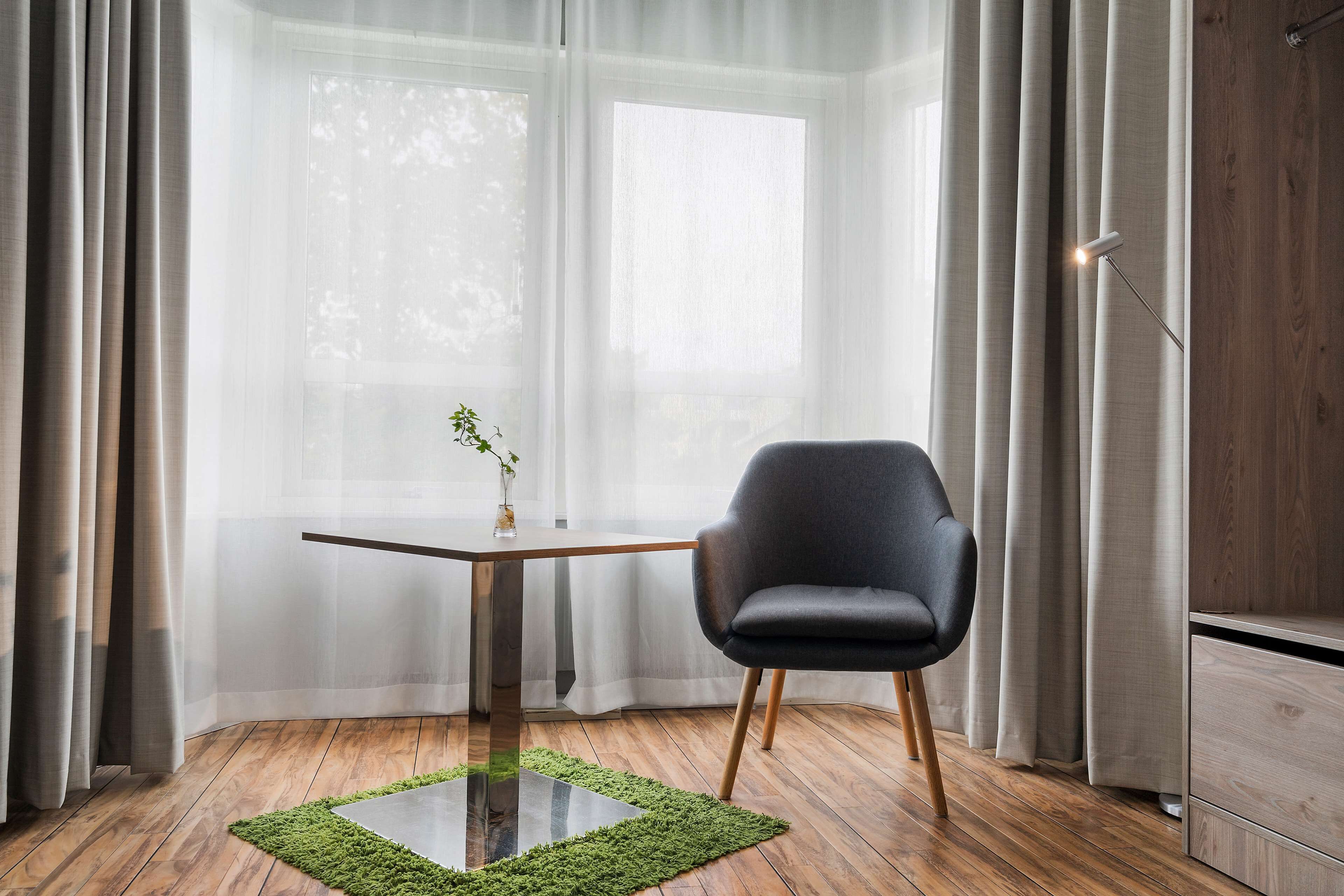 Une pièce dotée d’un plancher en bois, d’une table en verre surmontée d’un tapis vert, d’une chaise noire, d’un meuble en bois et de rideaux légers laissant passer la lumière naturelle.