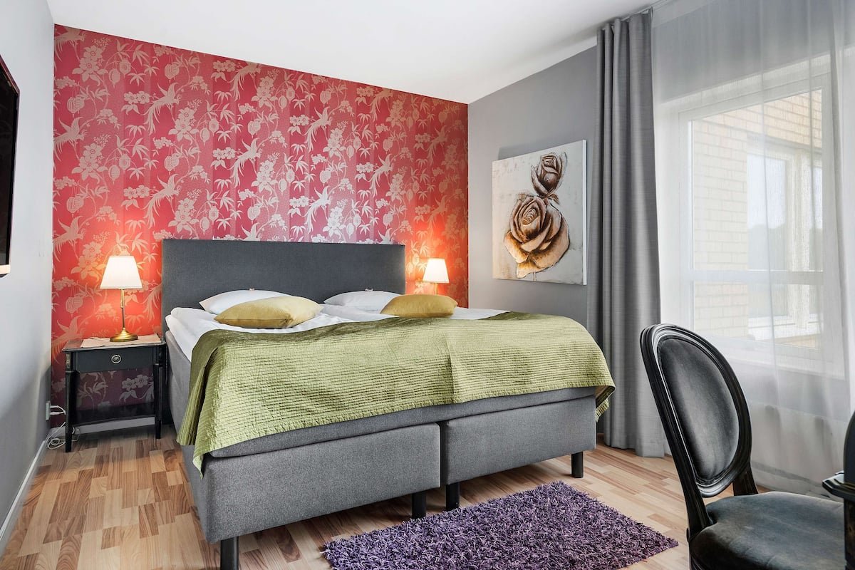 A bedroom with a bed, two bedside lamps, a purple rug, a chair, and a large window with curtains.