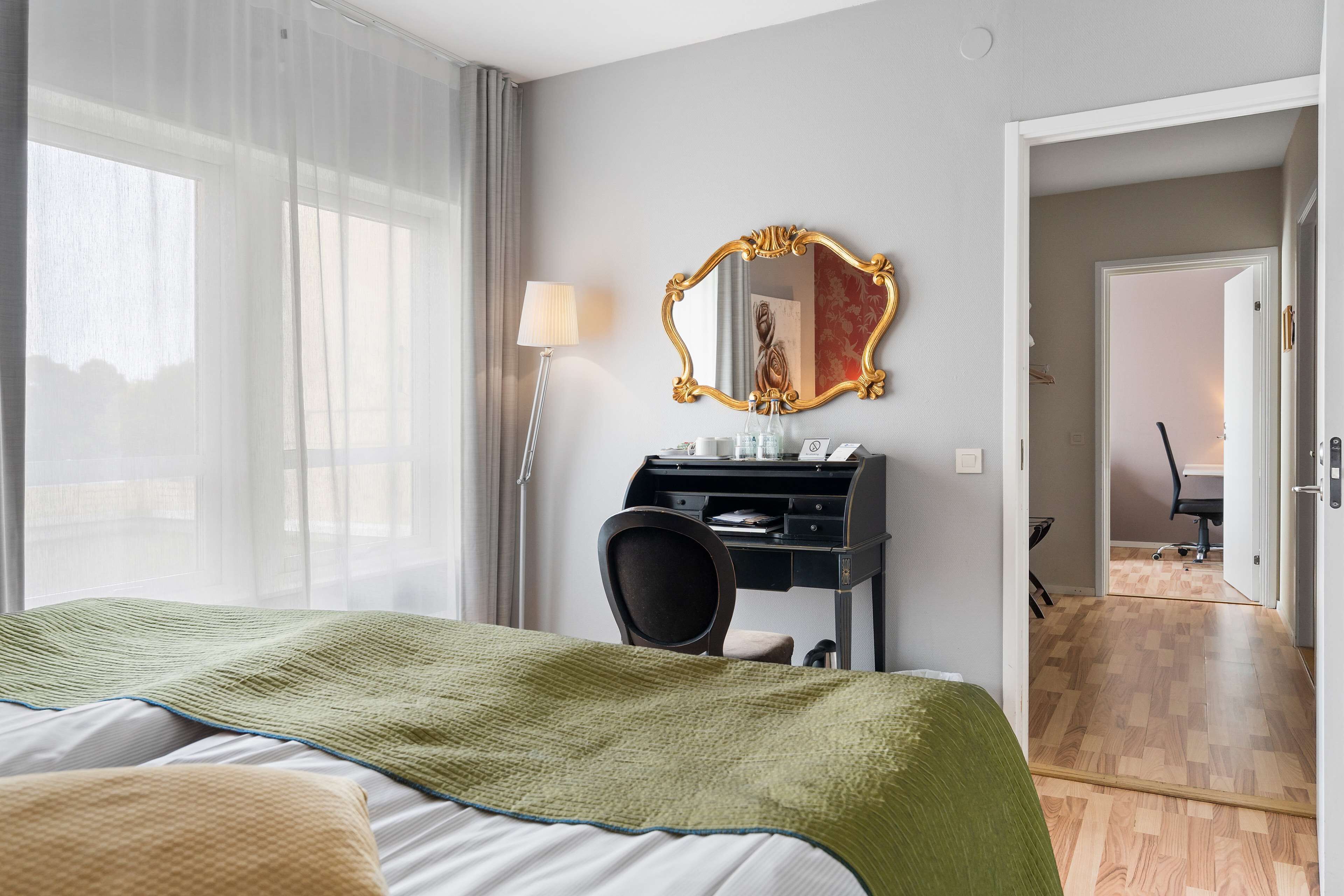 Une chambre d’hôtel avec un lit, un bureau avec une lampe, un miroir et une vue sur une autre chambre.